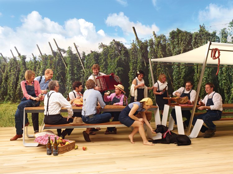 Gruppe genießt Picknick auf Extremis Hopper Picknickbank im Freien mit Speisen, Getränken und Akkordeonmusik vor Hopfenfeld-Hintergrund.