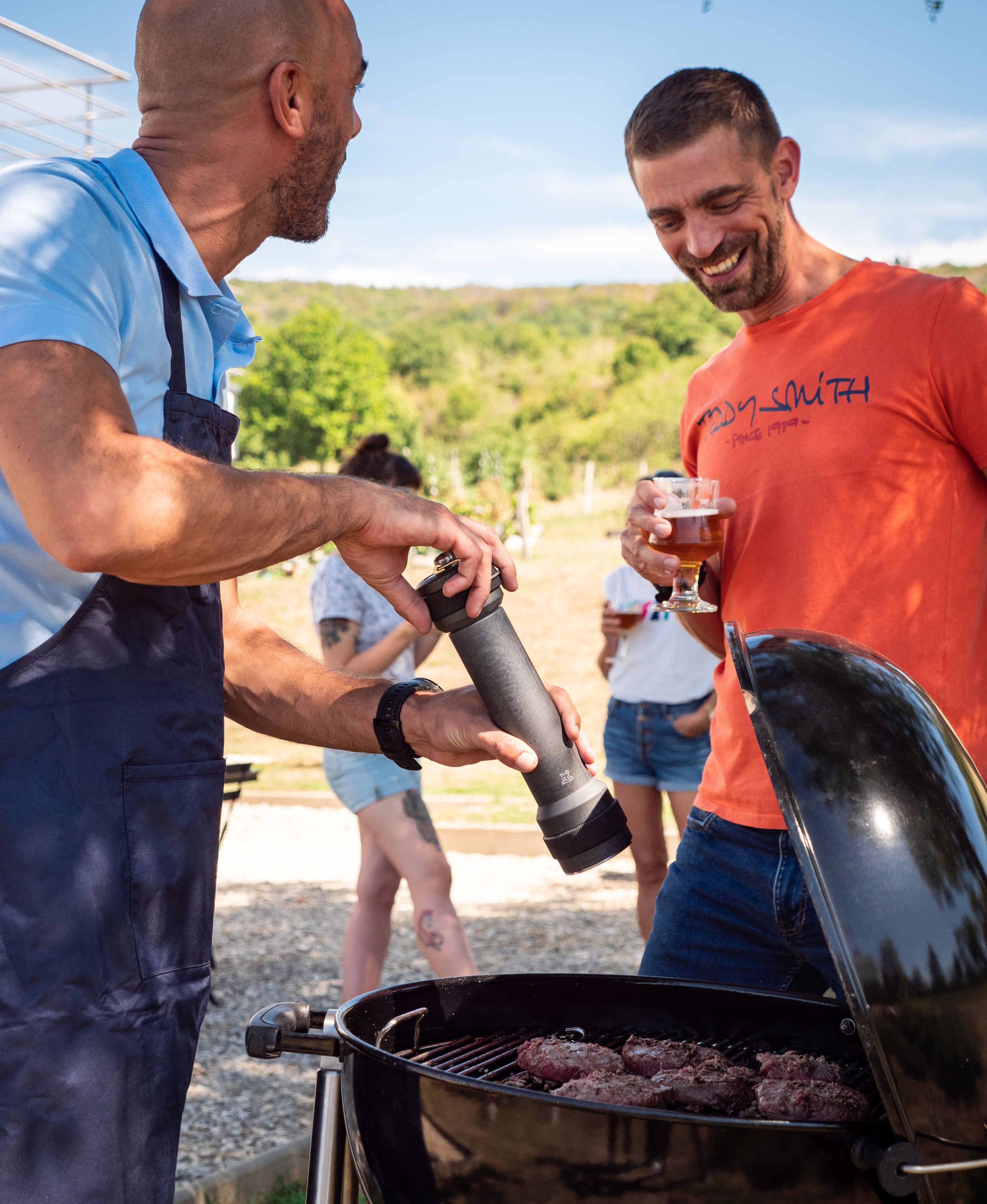 Mann würzt Burger auf Grill mit Peugeot BBQ Pfeffermühle, während Freund mit Bier zusieht.