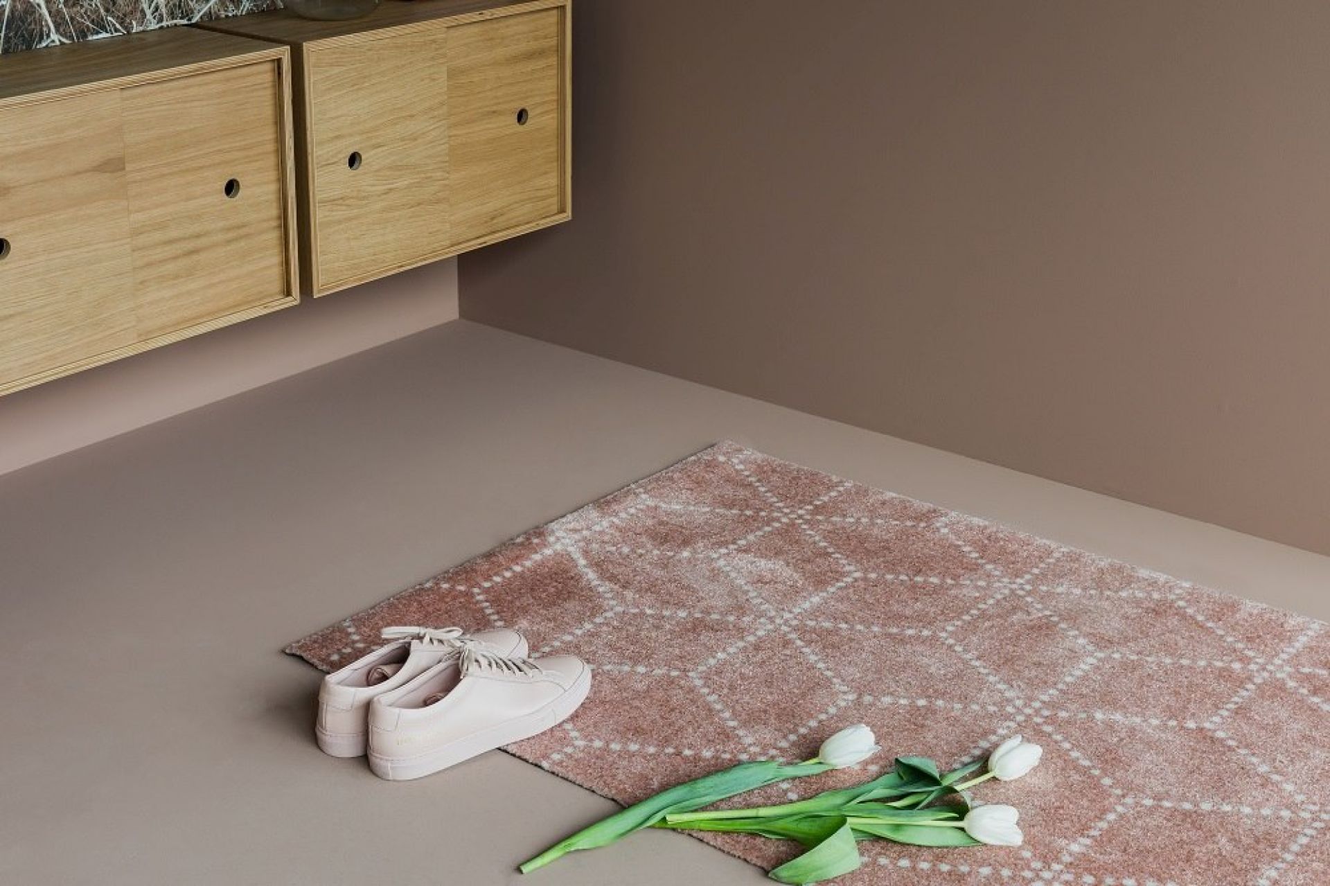 Rosa Teppich mit geometrischem Muster, weißen Schuhen und Tulpen vor Holzschrank und brauner Wand.