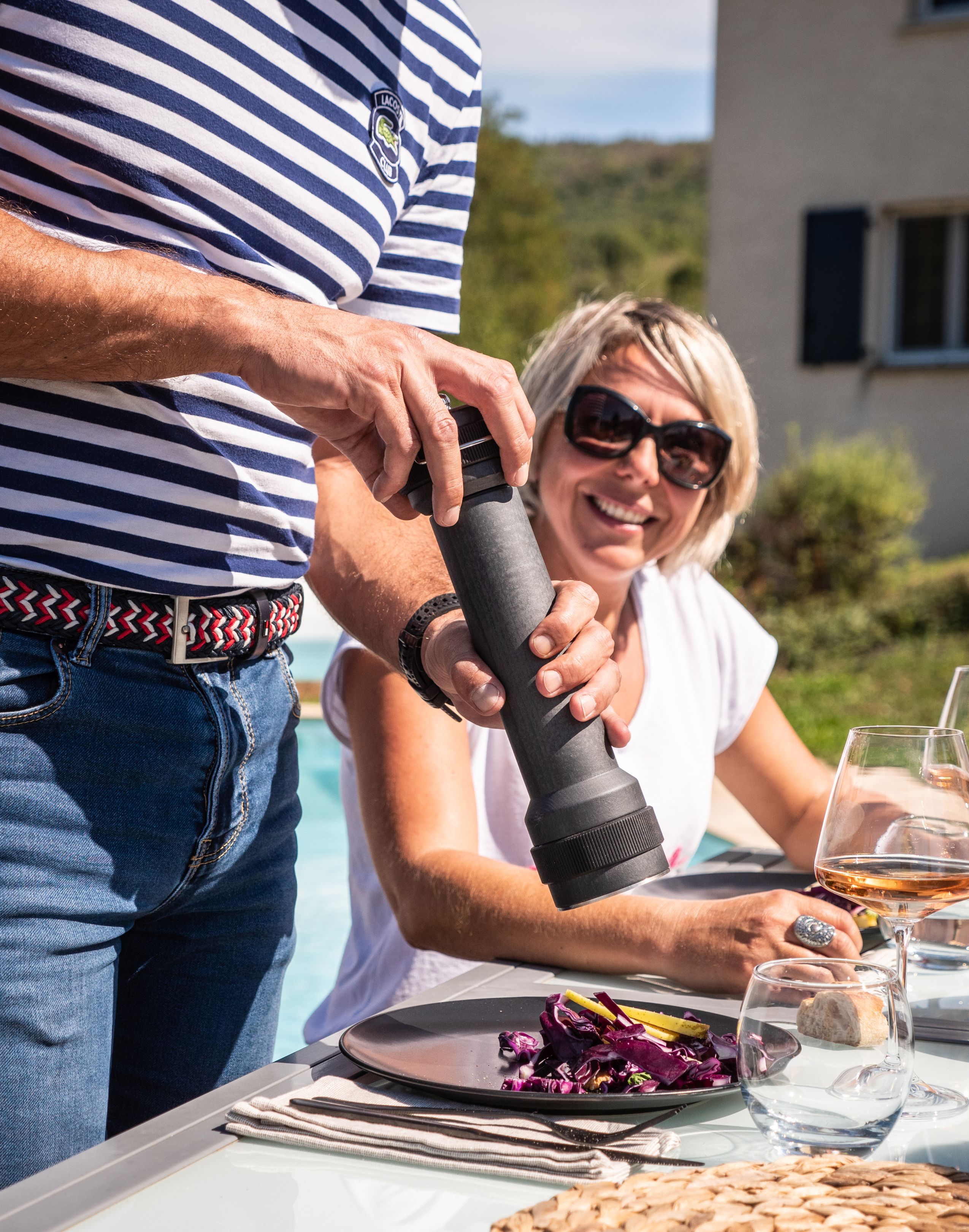 Mann würzt Salat mit Peugeot BBQ Pfeffermühle im Freien, Frau lächelt im Hintergrund.