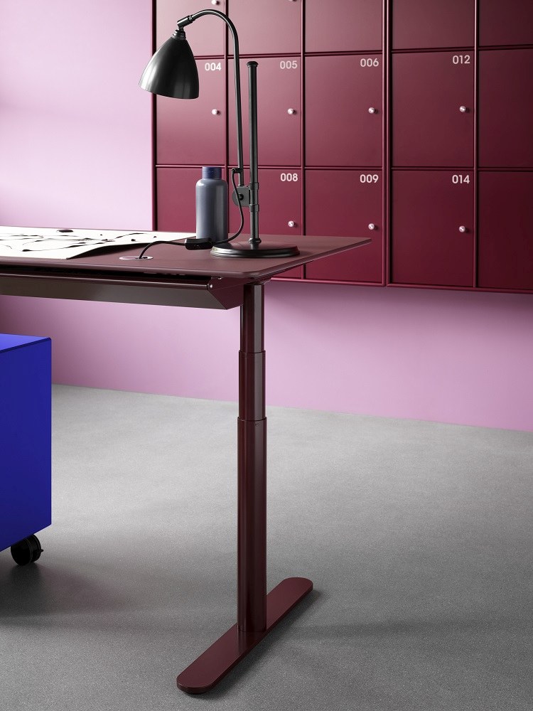 HiLow 2 höhenverstellbarer Schreibtisch von Montana in Bordeauxrot mit Lampe und Rollcontainer vor Spindwand.