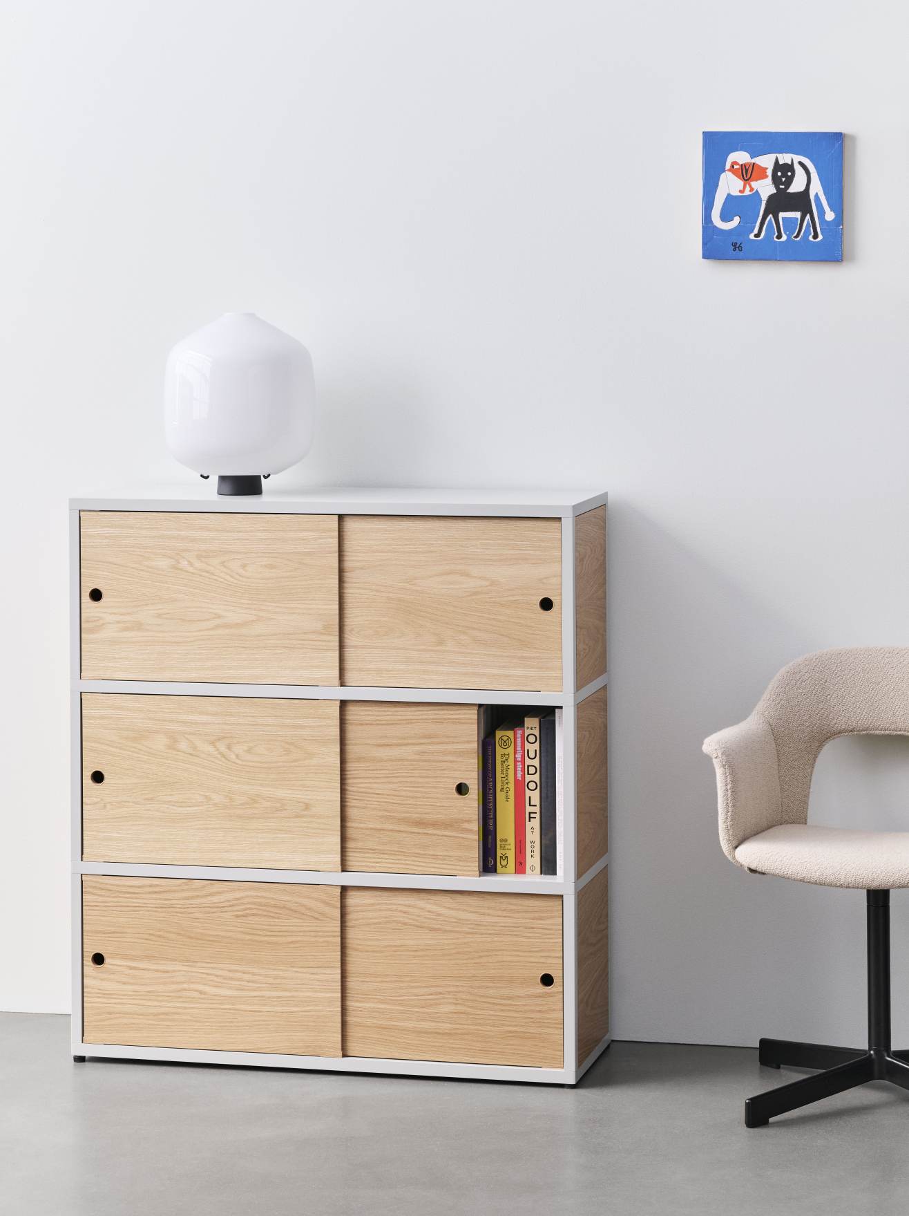 Heller Raum mit Hay Armlehnstuhl, Bücherregal mit Schiebetüren, Lampe und Wandbild.