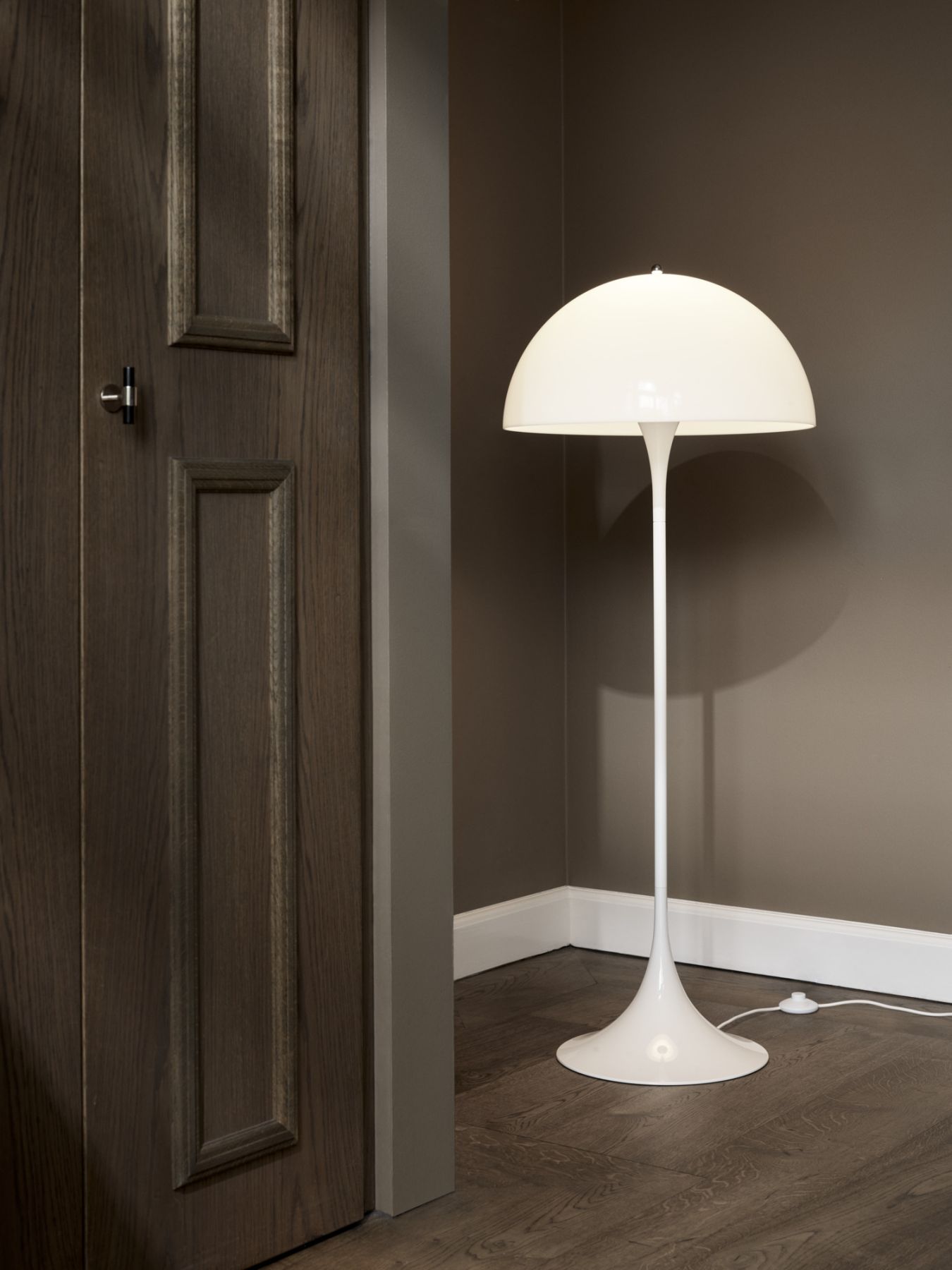 Moderne weiße Stehlampe im Pilzdesign neben einer dunklen Holztür auf dunklem Holzboden.