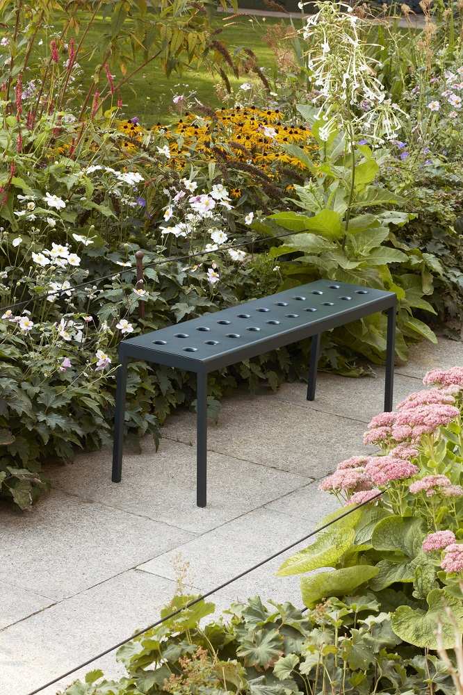 Hay Balcony Bank für den Außenbereich, kleine graue Gartenbank mit Löchern im Sitz, umgeben von Blumen.