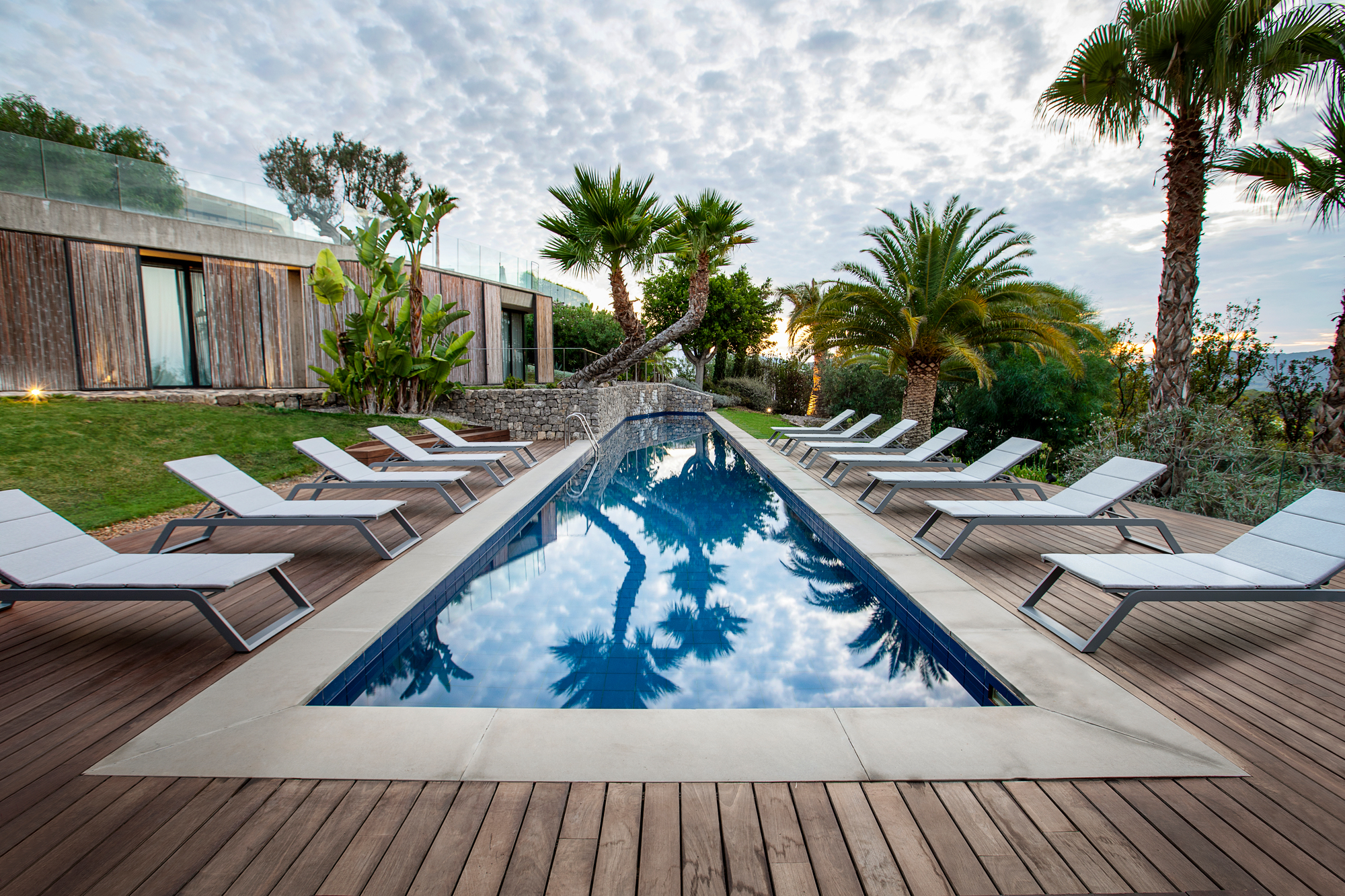 Cane-Line Siesta Sonnenliegen aus grauem Aluminium und Textil am Pool auf Holzdeck mit Palmen.