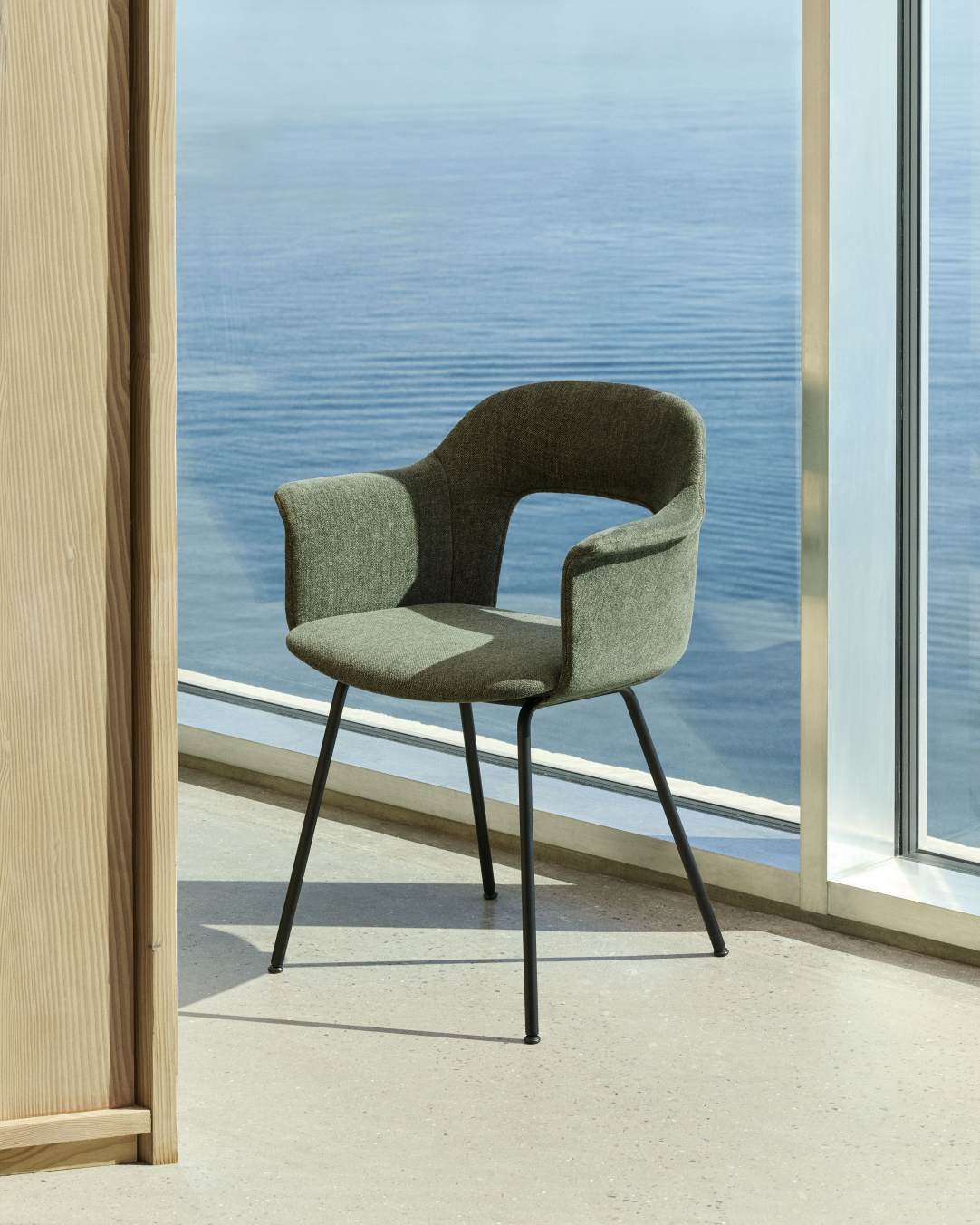 Layout Armchair von Hay, grüner Armlehnstuhl mit schwarzem Metallgestell vor einem Fenster mit Meerblick.
