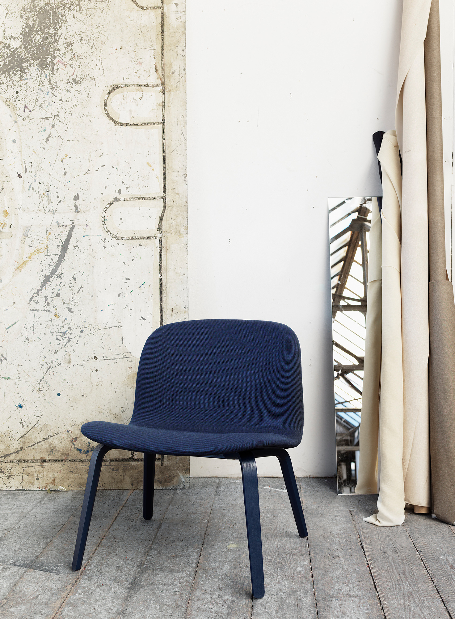 Blauer Visu Lounge Chair von Muuto vor einer Wand mit Spiegel und Stoffrollen.