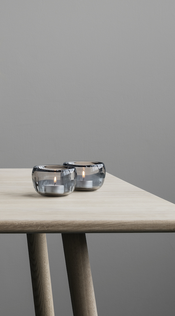 Zwei graue Stelton Ora Teelichthalter aus Glas mit brennenden Teelichtern auf einem Holztisch.