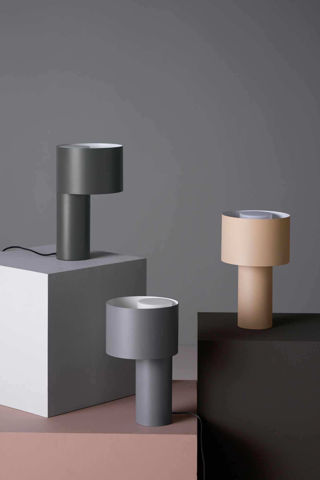 Drei moderne Tangent Tischleuchten von Woud in Grau, Beige und Olivgrün auf geometrischen Blöcken.