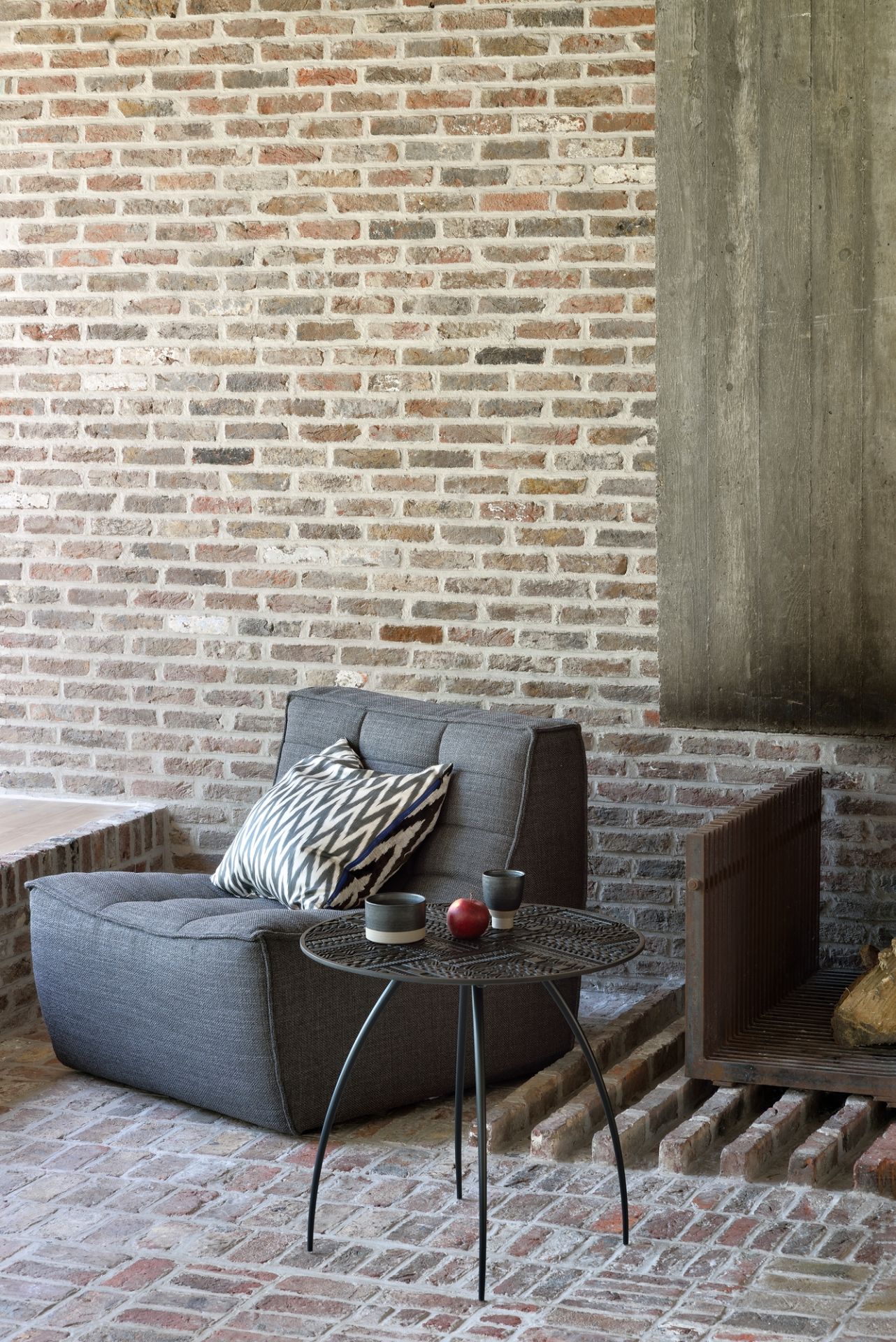 Grauer Loungesessel mit Kissen und kleinem Beistelltisch auf Backsteinboden, modernes Wohnzimmer Interieur.