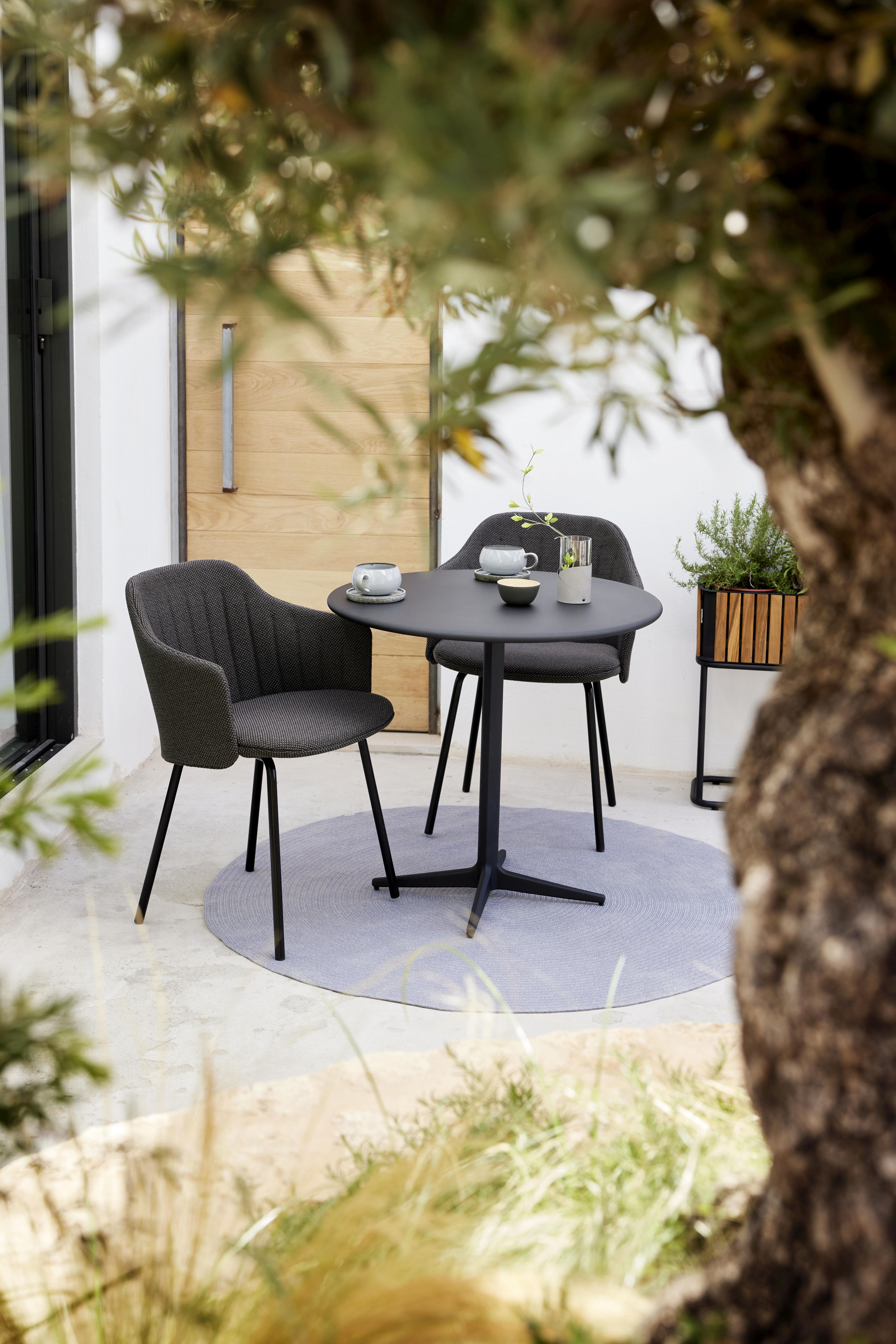 Outdoor-Szene mit Cane-Line Stühlen und rundem Tisch auf einem taupefarbenen Teppich.