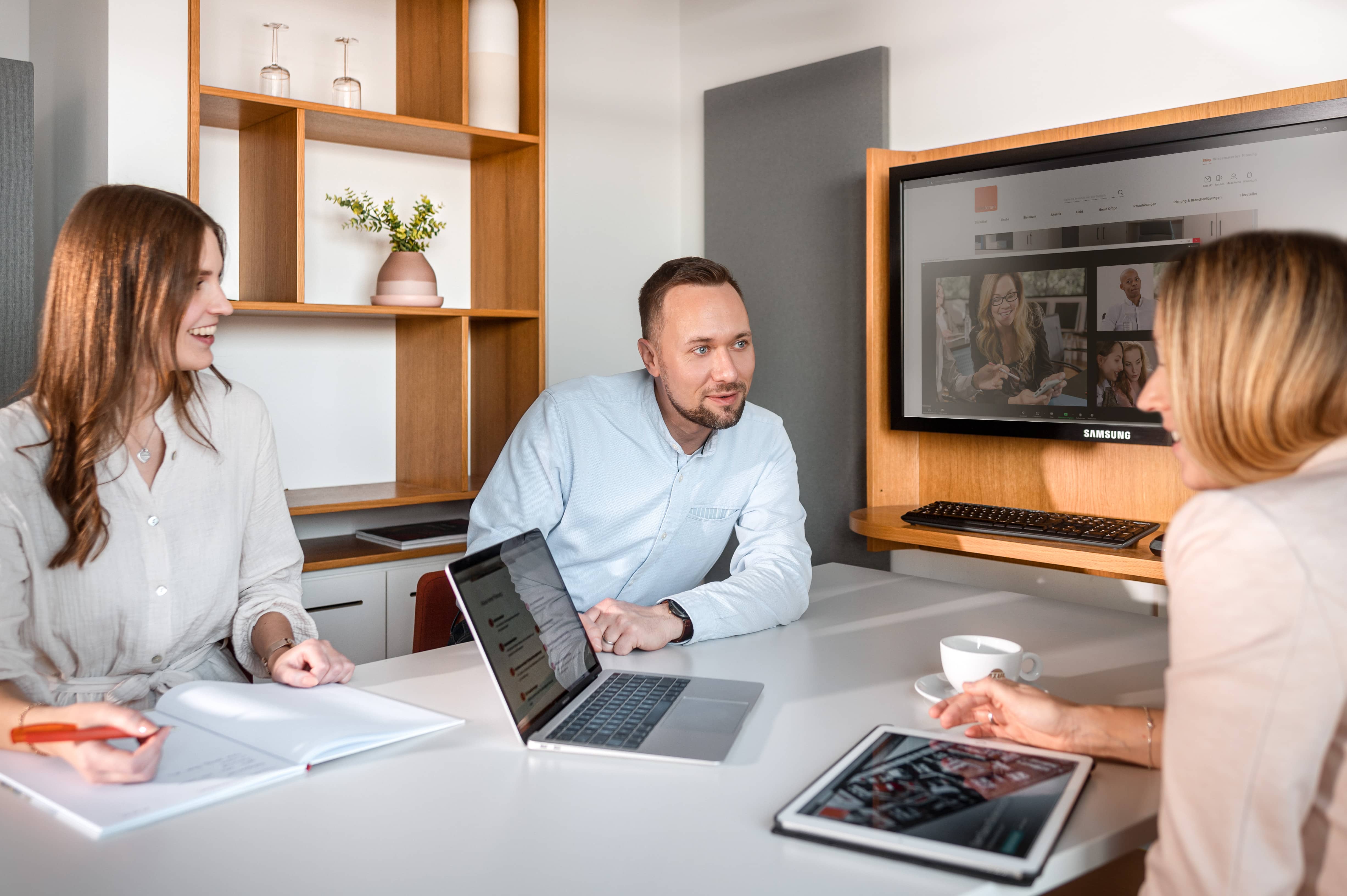 Drei Personen im Büro bei einer Videokonferenz mit Laptop, Tablet und großem Bildschirm für virtuelle Meetings.