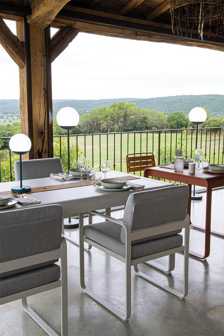Bellevie Sessel von Fermob, graue Outdoor-Sitzmöbel mit Tisch und Beleuchtung auf einer Terrasse mit Ausblick.