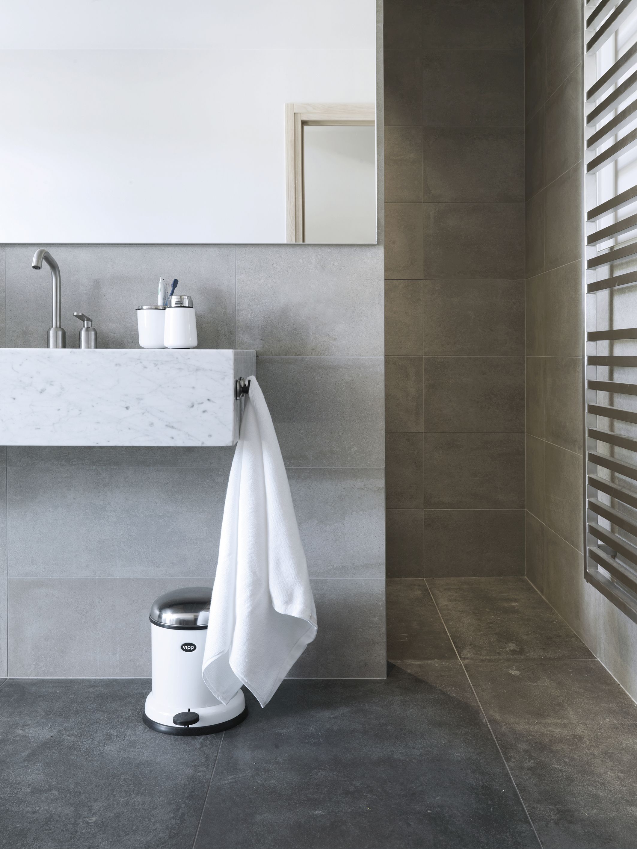 Weißer Vipp Treteimer aus Stahl im modernen Badezimmer mit Handtuch und grauen Fliesen.