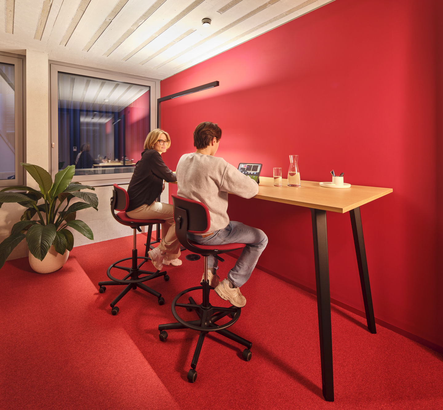 Zwei Personen sitzen auf roten Vitra Rookie High Bürodrehstühlen an einem Stehtisch in einem modernen Büro.