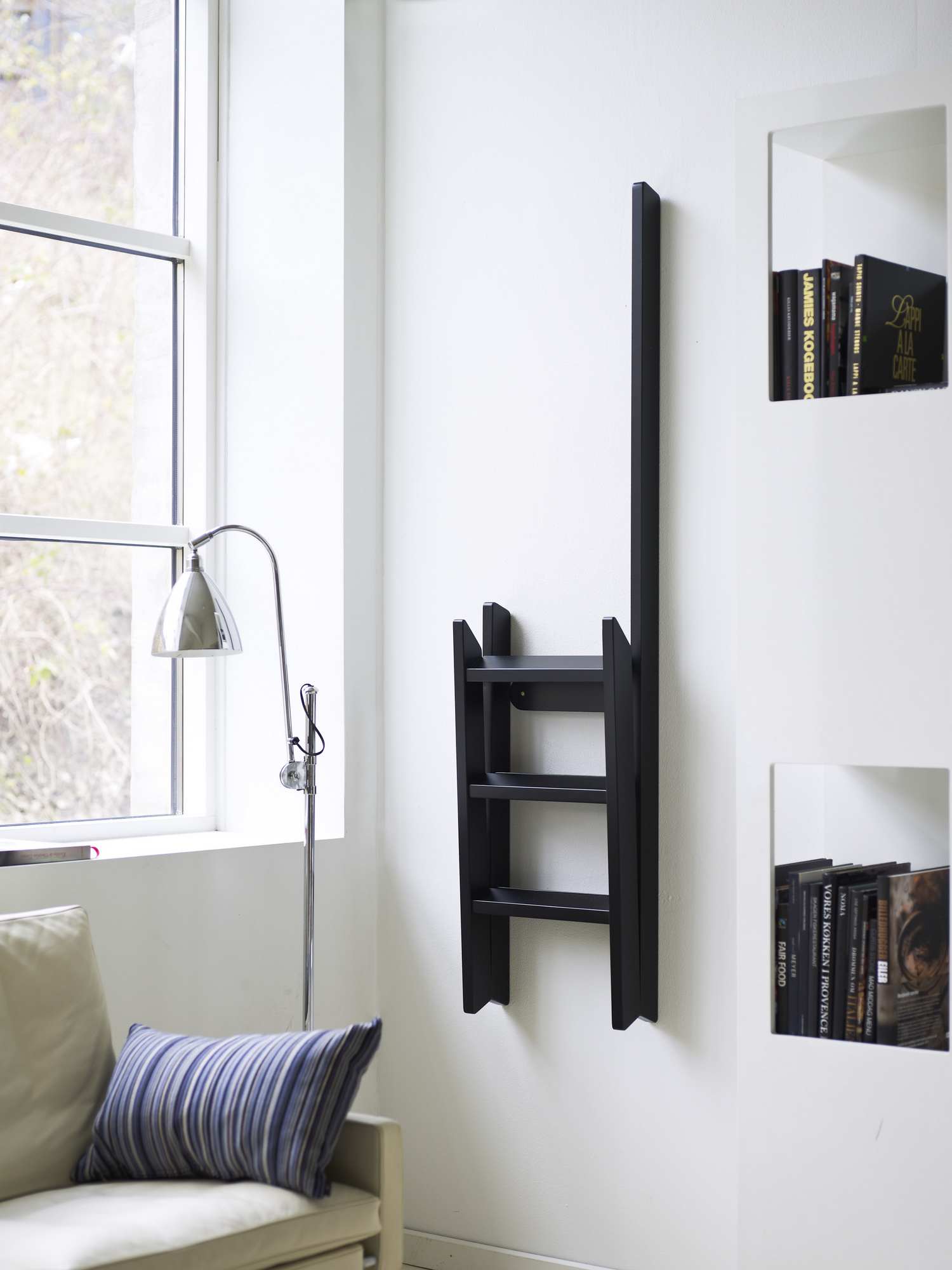 Schwarze Hindevadgaard Trittleiter an weißer Wand, zusammengeklappt als Dekoelement, modernes Wohnzimmer mit Fenster und Lampe.