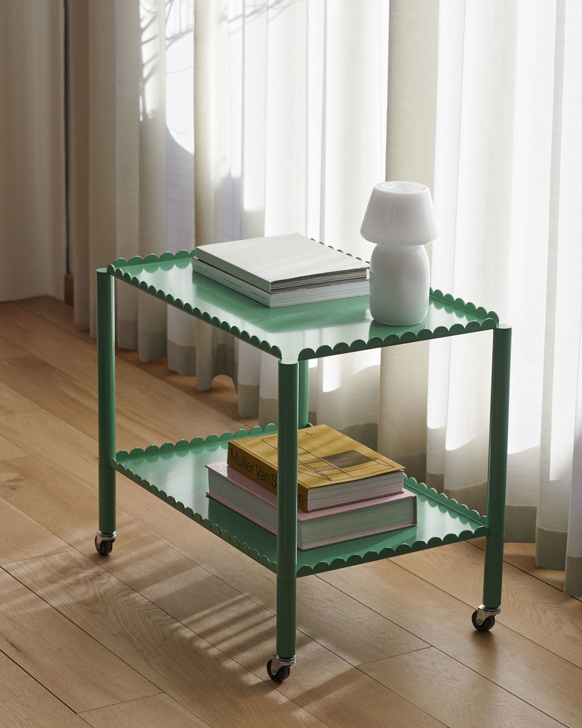 Grüner Arcs Trolley Servierwagen von Hay mit zwei Ablagen und Büchern vor hellem Vorhang.