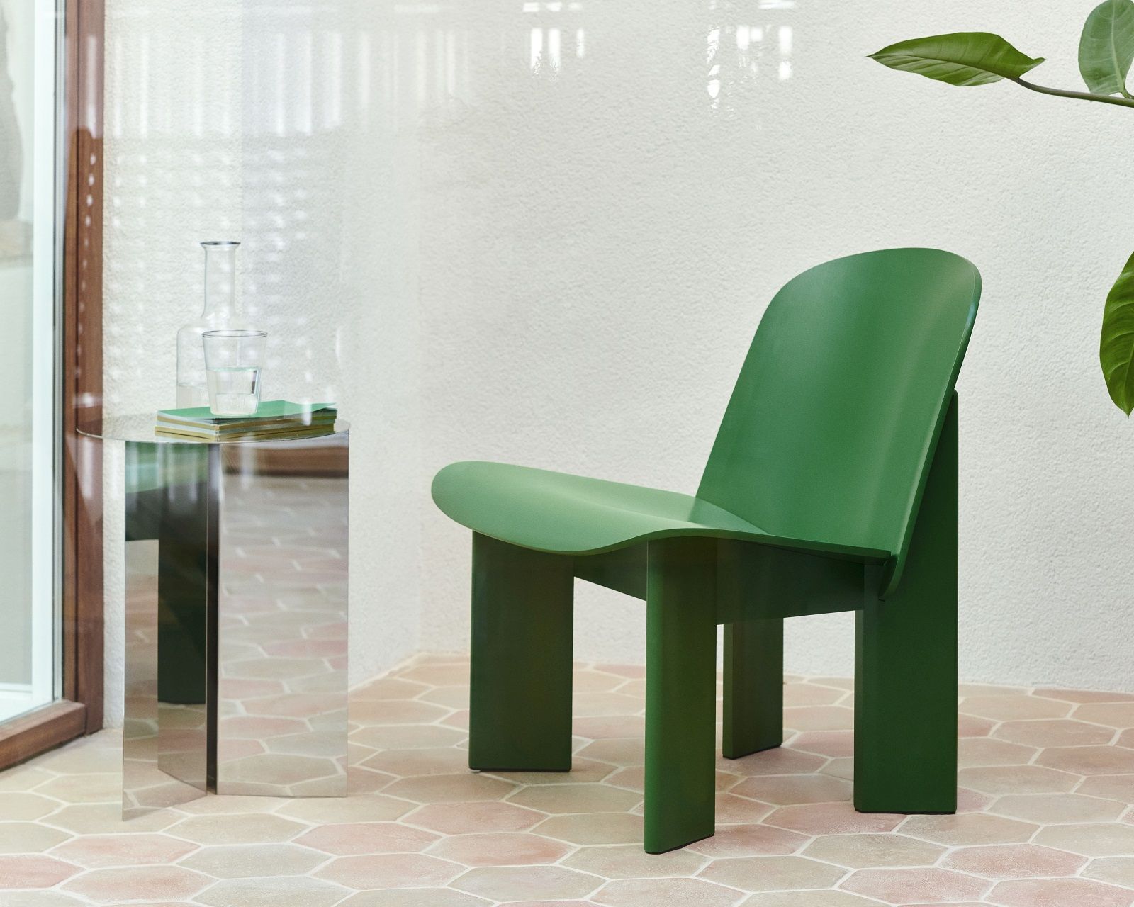 Hay Slit Table Round High Beistelltisch aus Spiegelmetall mit Wasserflasche und Glas, daneben grüner Stuhl.
