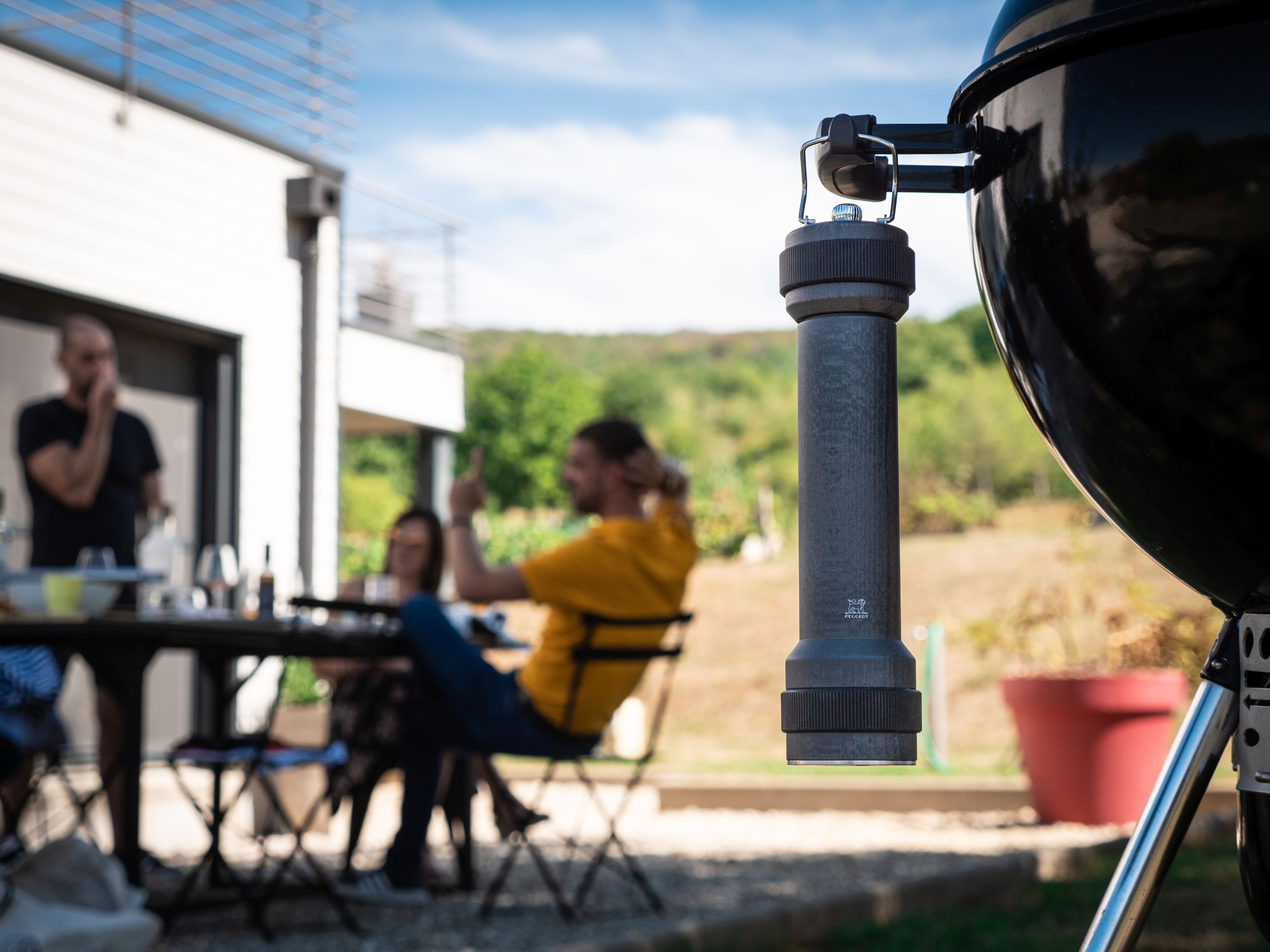 Graphitfarbene Peugeot BBQ Pfeffermühle am Grill befestigt, im Hintergrund eine entspannte Grillrunde im Garten.