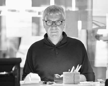 Schwarz-Weiß-Porträt eines Mannes mit Brille und dunklem Pullover am Schreibtisch im Büro.