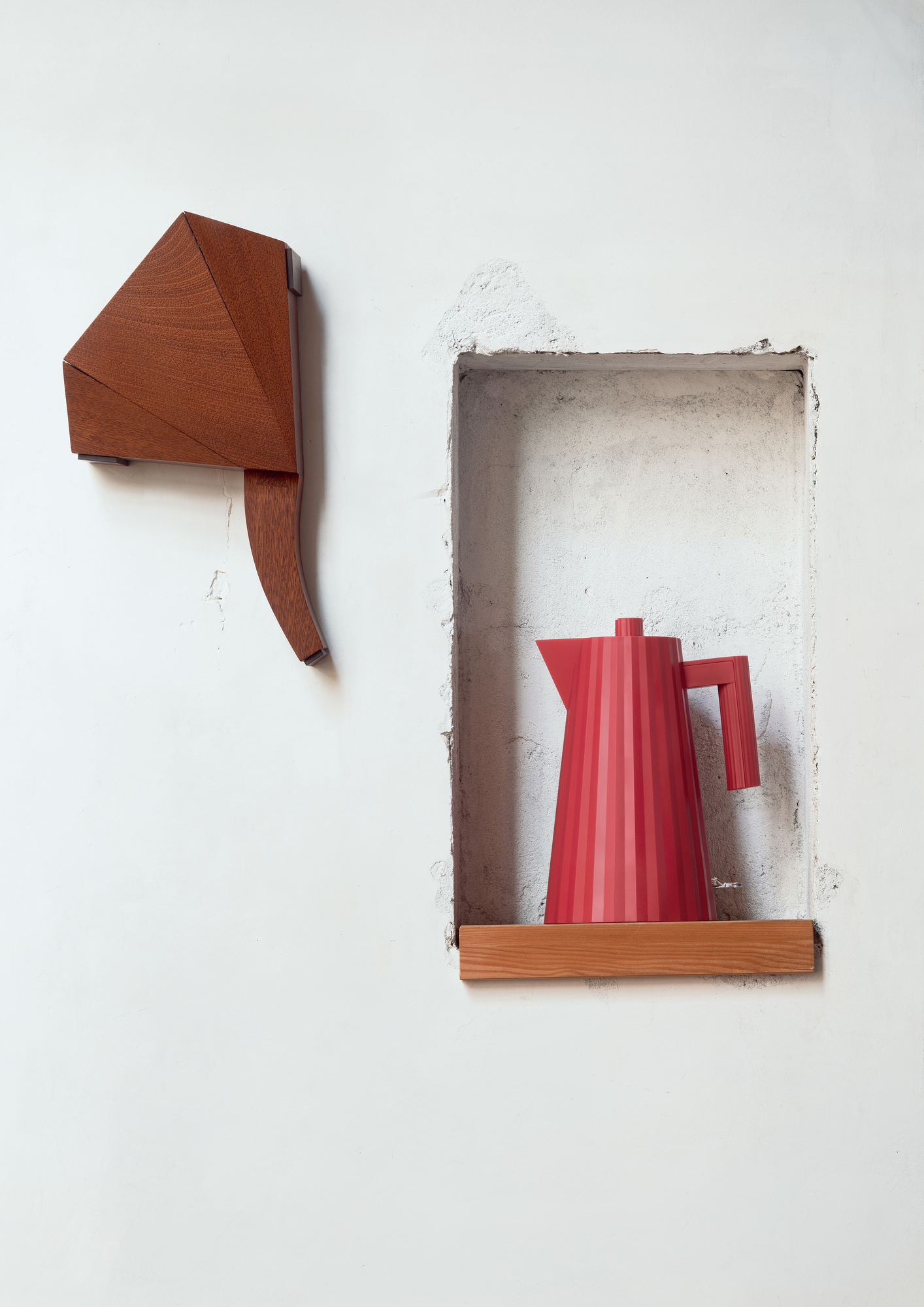 Roter Alessi Plissé Wasserkocher in Nische, daneben Holzobjekt an weißer Wand. Design Wasserkocher.