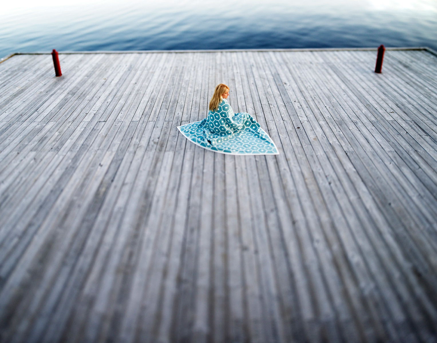 Frau sitzt auf Holzsteg, gehüllt in eine blaue Pappelina Wolldecke mit Muster.