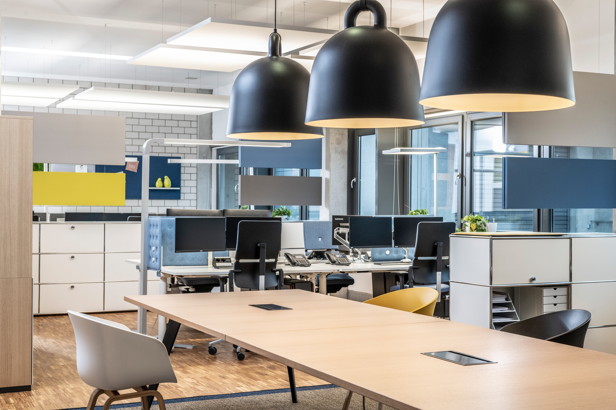 Helle Büroraum-Szene mit drei schwarzen Normann Copenhagen Bell Lamp Hängeleuchten über einem langen Holztisch.
