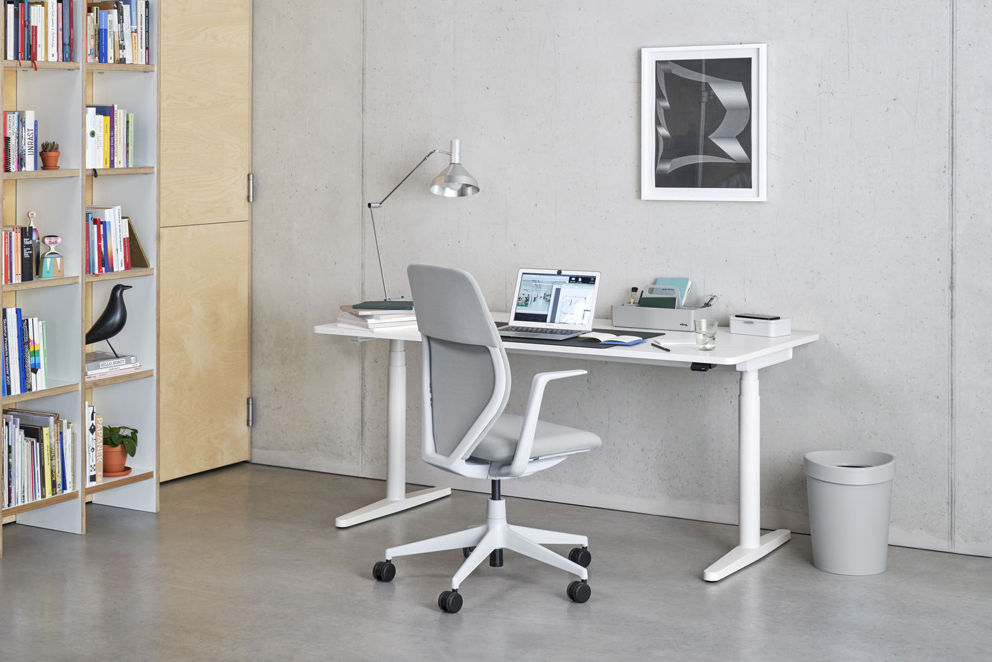 Heller Arbeitsplatz mit Vitra Tyde 2 Steh-Sitz-Schreibtisch, Bürostuhl, Lampe und Laptop vor einer Betonwand.