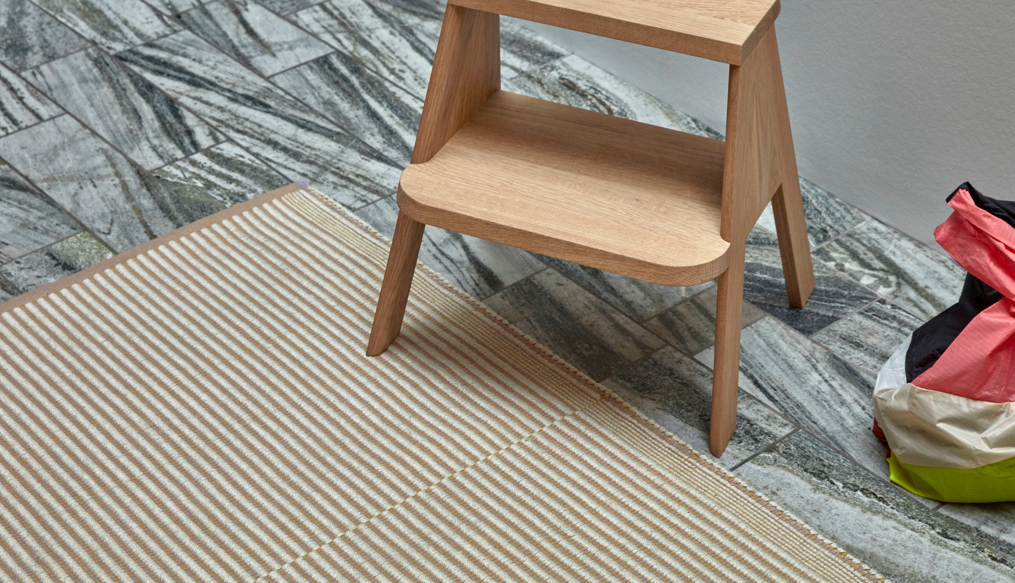 Detailaufnahme des Hay Tapis Teppichs mit beigen Streifen und einem Holzstuhl auf grauem Fliesenboden.