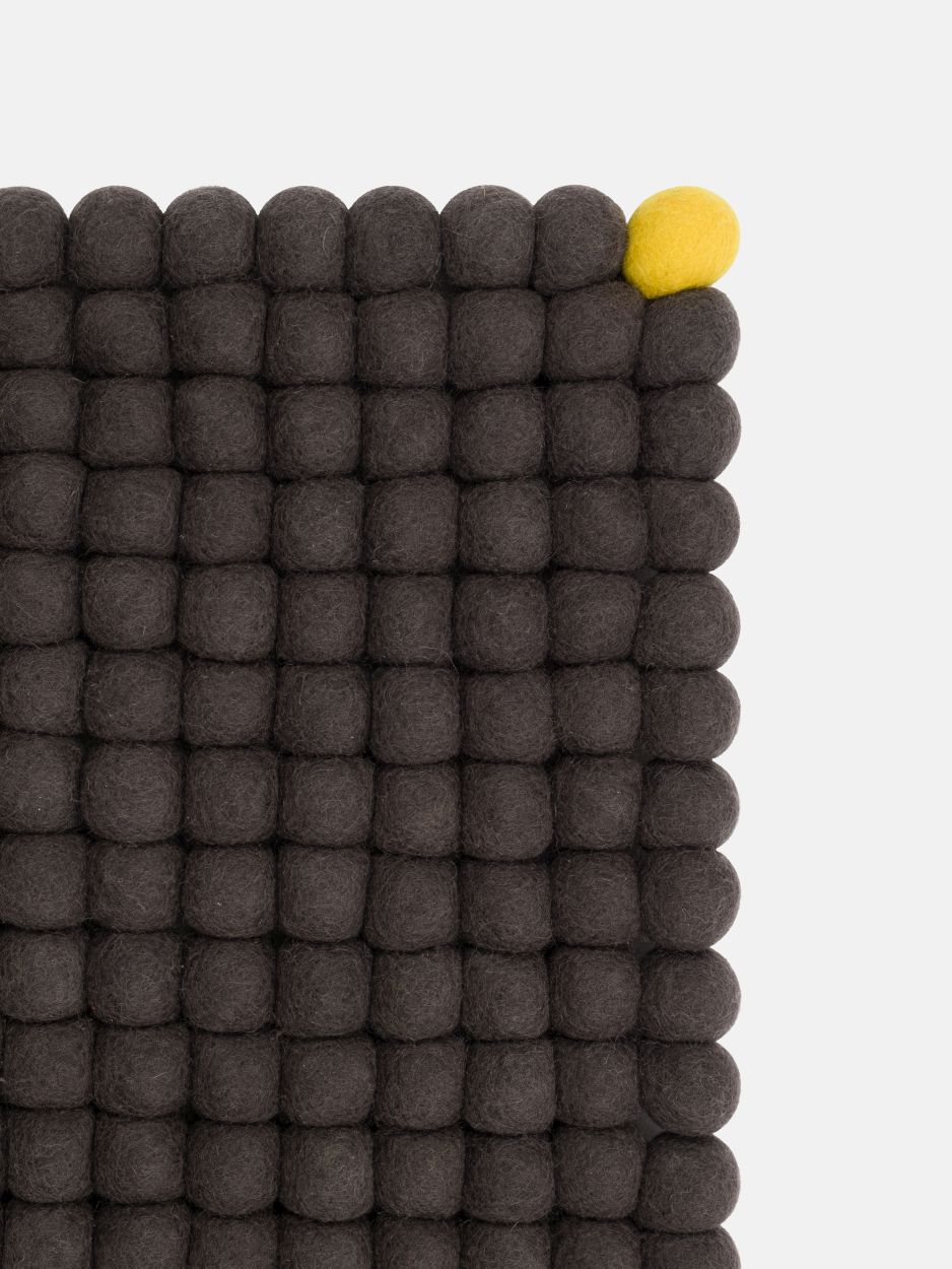 Detailaufnahme: Anthrazitfarbener Filzteppich mit Filzkugeln, ein gelber Akzent. Myfelt Teppich für Wohnräume.
