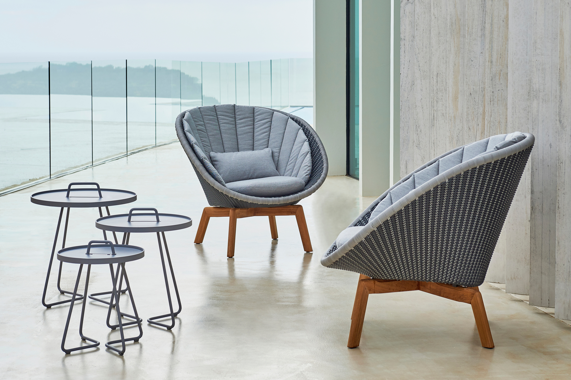 Zwei graue Cane-Line Peacock Loungesessel aus Rattan mit Kissen und drei graue Beistelltische im Außenbereich.