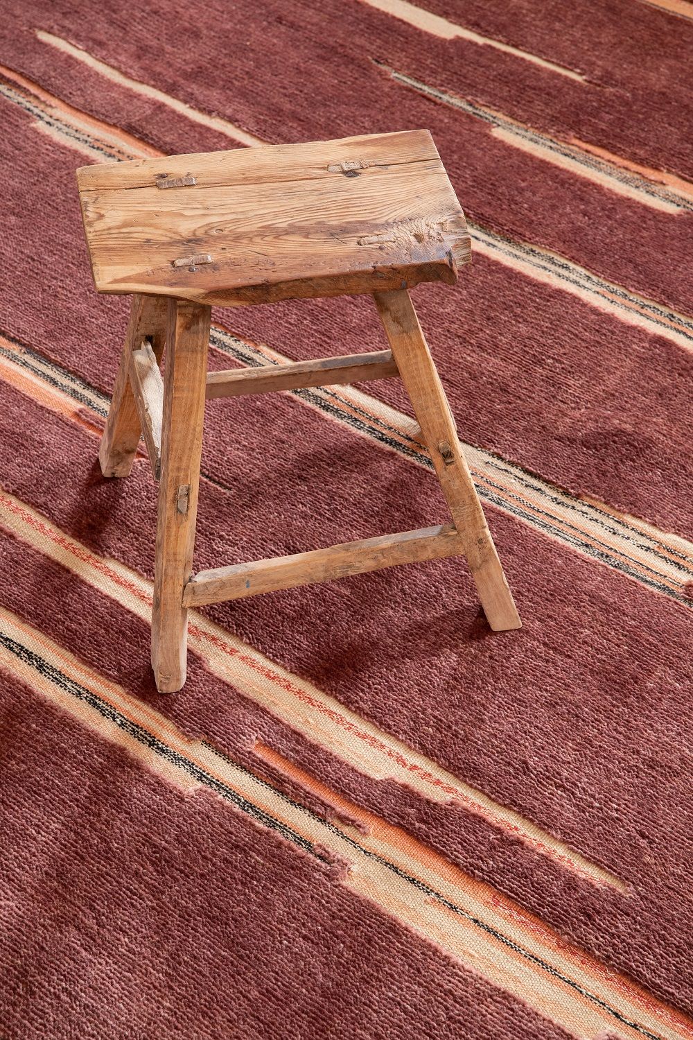 Colorado Teppich von Nanimarquina, Detailaufnahme mit Holzschemel auf dem gestreiften, bordeauxroten Wollteppich.