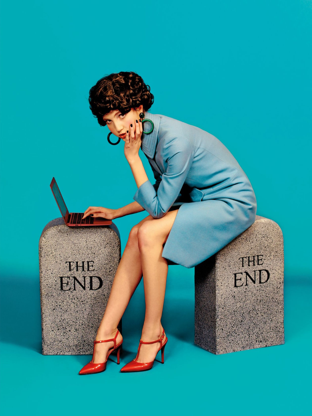 Frau im blauen Kleid, rote High Heels, sitzt auf Grabsteinen mit Laptop. Modische Accessoires.