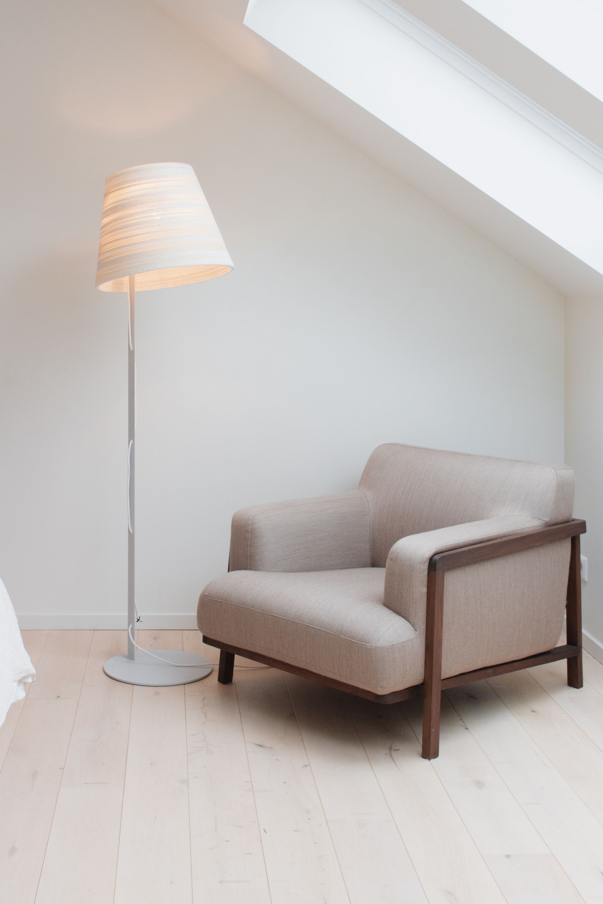 Weiße Tilt Stehleuchte Scraplights von Graypants neben beigem Sessel in hellem Raum mit Dachfenster.
