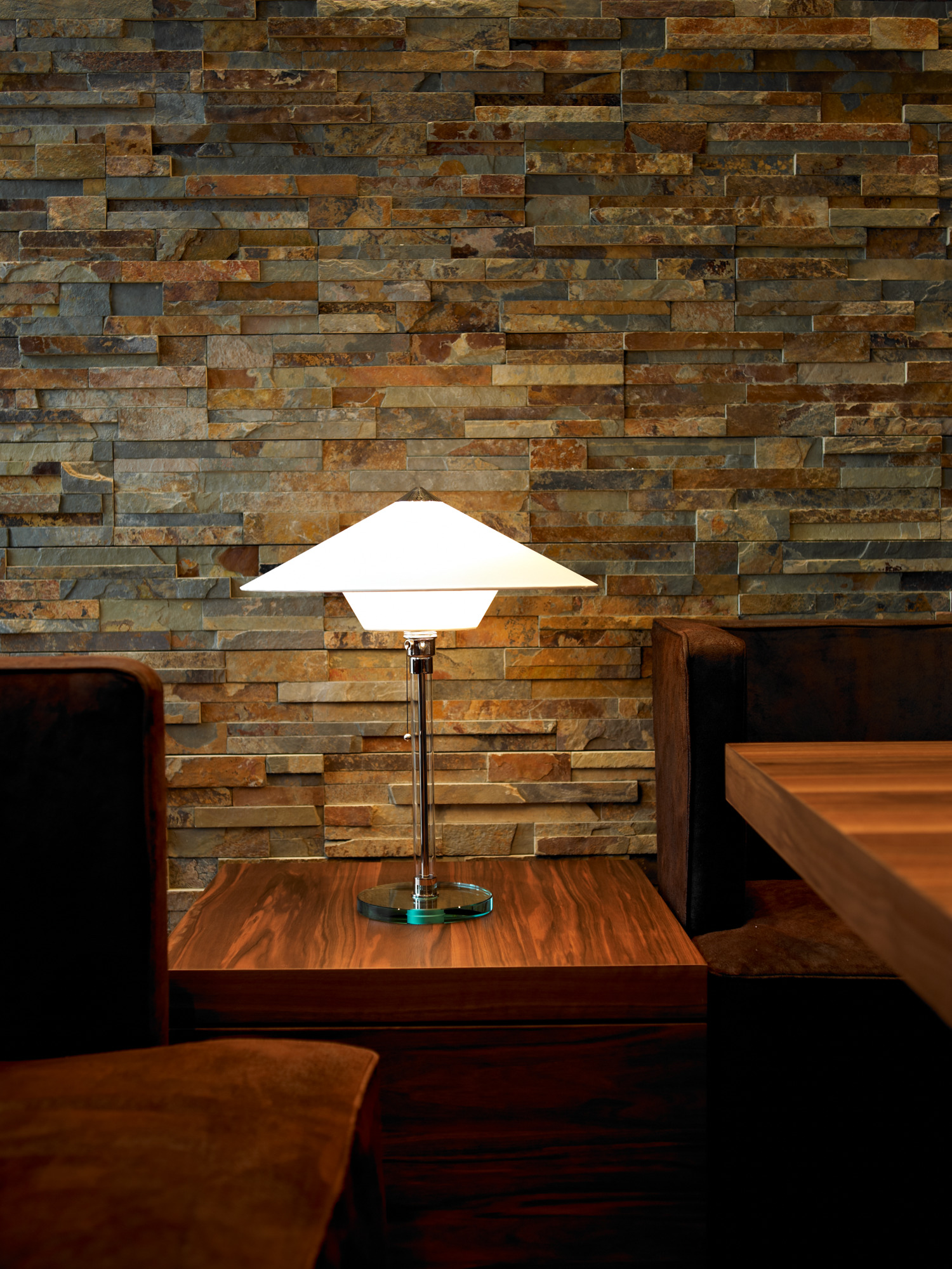 Wagenfeld Tischleuchte WG28 von Tecnolumen auf dunklem Holztisch vor einer Steinwand. Designklassiker Bauhaus-Stil.