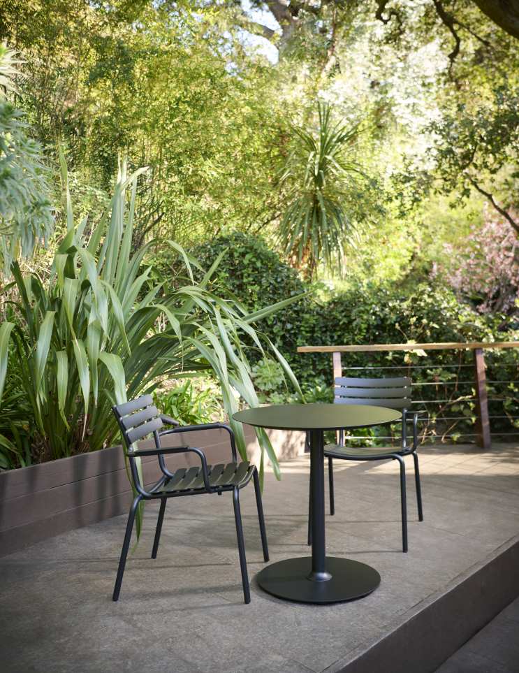 Alua Armlehnenstuhl von Houe mit passendem Gartentisch auf einer Terrasse im Grünen.