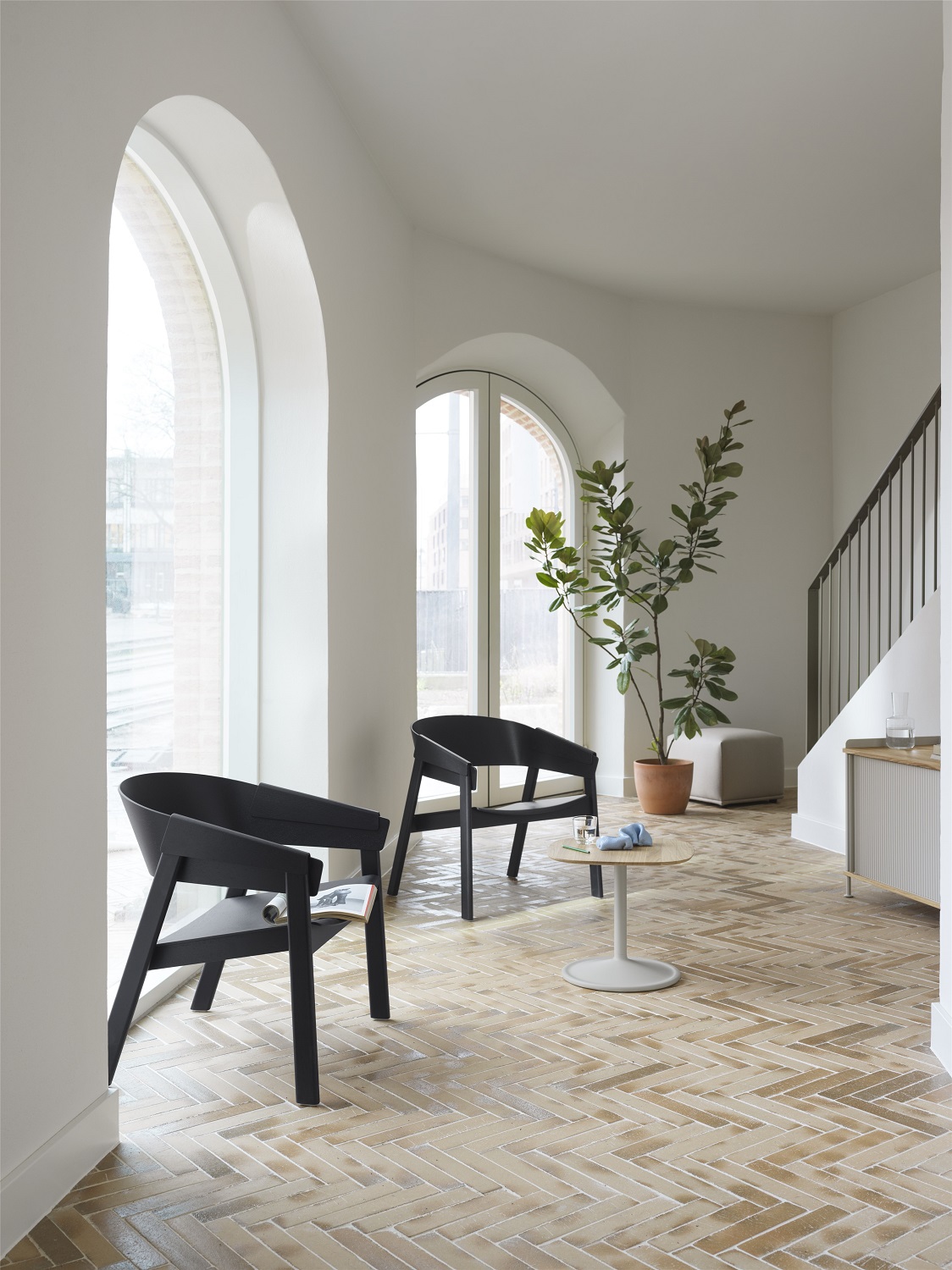 Elegantes Interieur mit zwei schwarzen Muuto Sesseln, kleinem Tisch und Pflanze, im Hintergrund ein Echo Pouf Hocker.
