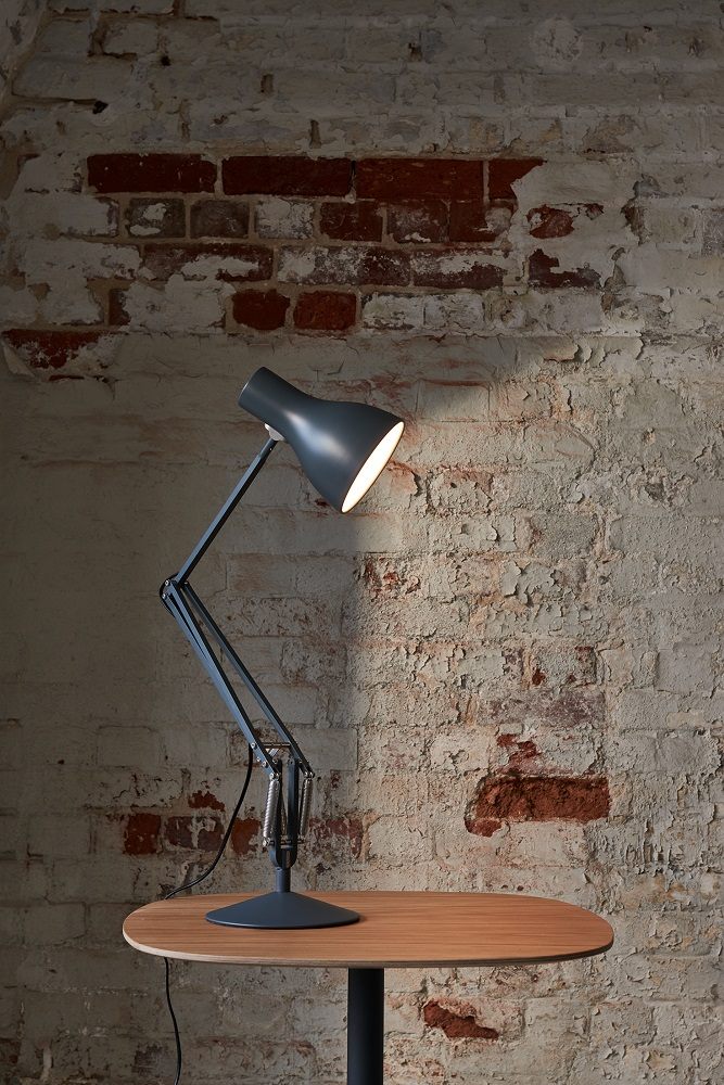 Graue Type 75 Schreibtischleuchte von Anglepoise auf einem Holztisch vor einer Backsteinwand.