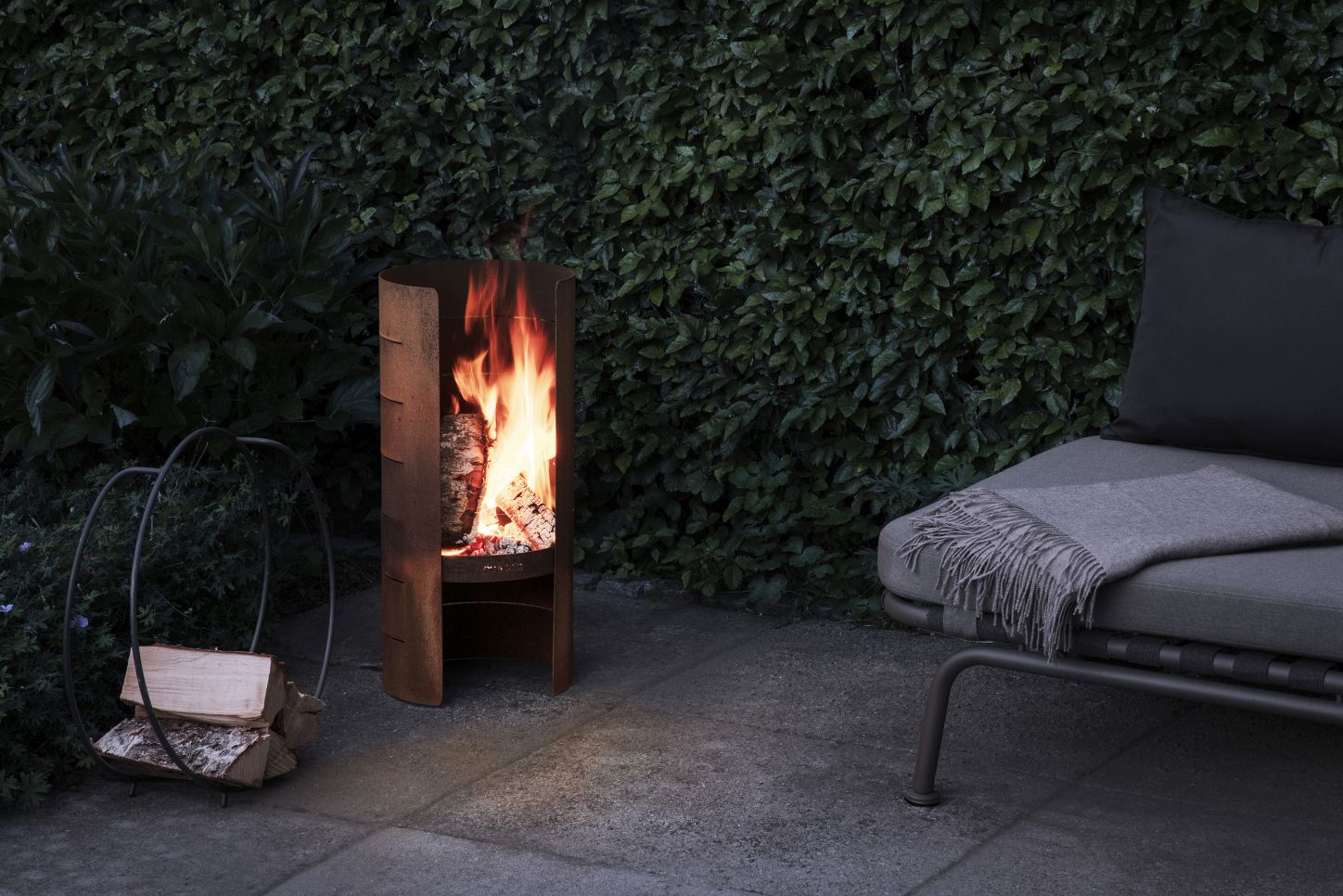 Eva Solo FireCylinder Gartenkamin aus Stahl mit brennendem Holz und daneben ein Holzkorb im Garten.