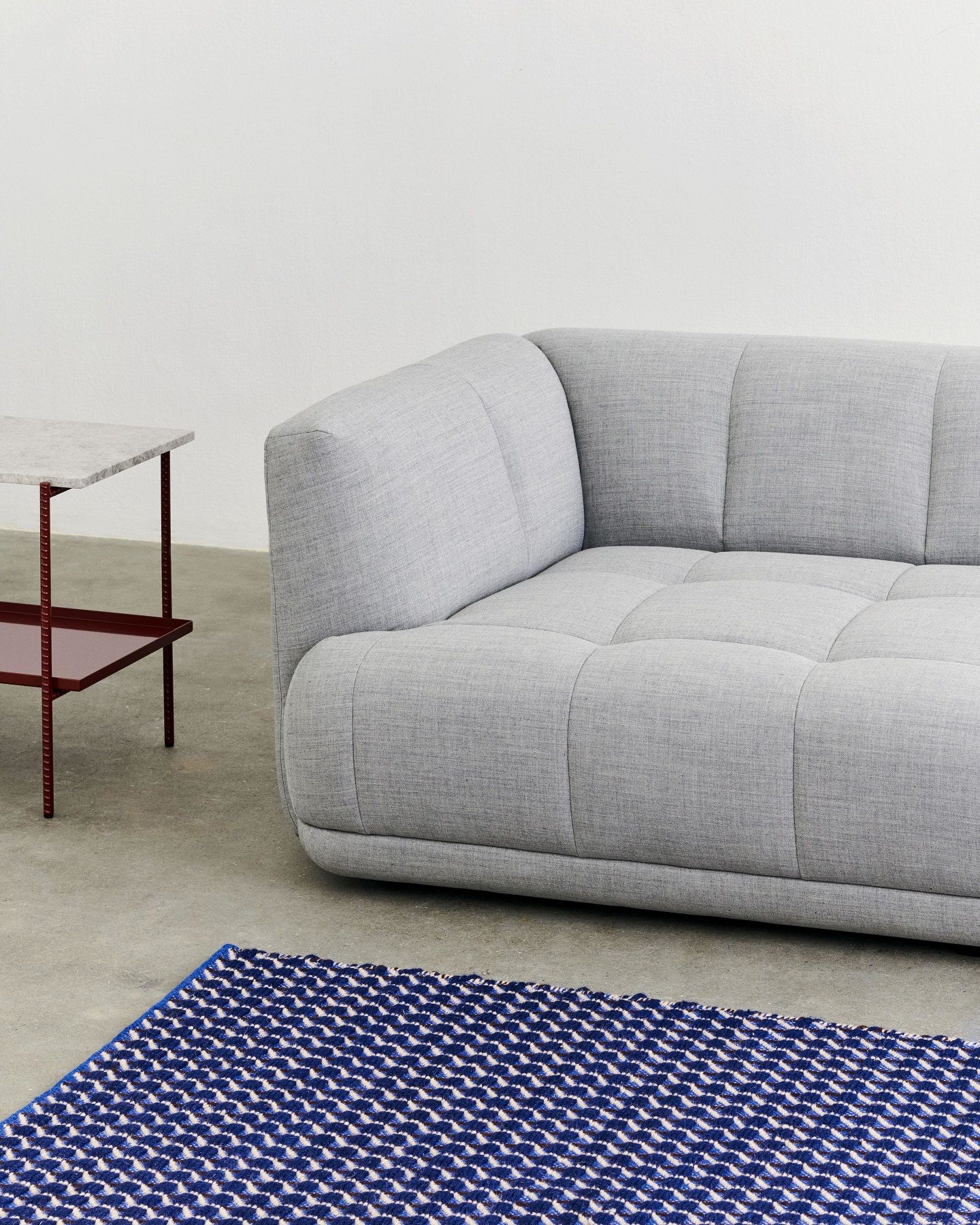 Nahaufnahme des Channel Rug Teppichs von Hay in Blau-Weiß mit geometrischem Muster, neben grauem Sofa.
