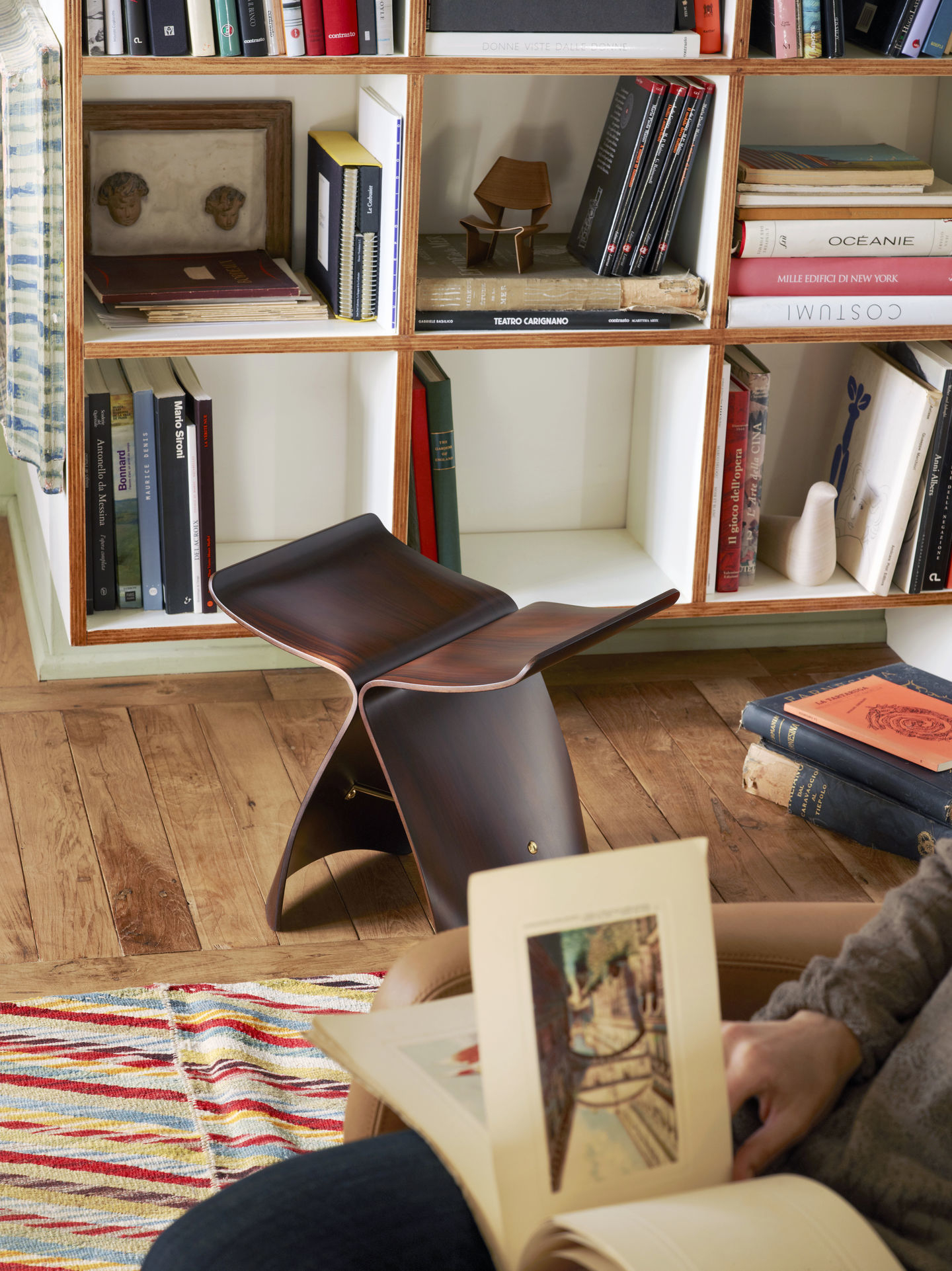 Vitra Butterfly Stool Hocker aus Walnussholz vor Bücherregal und Teppich im Wohnzimmer.