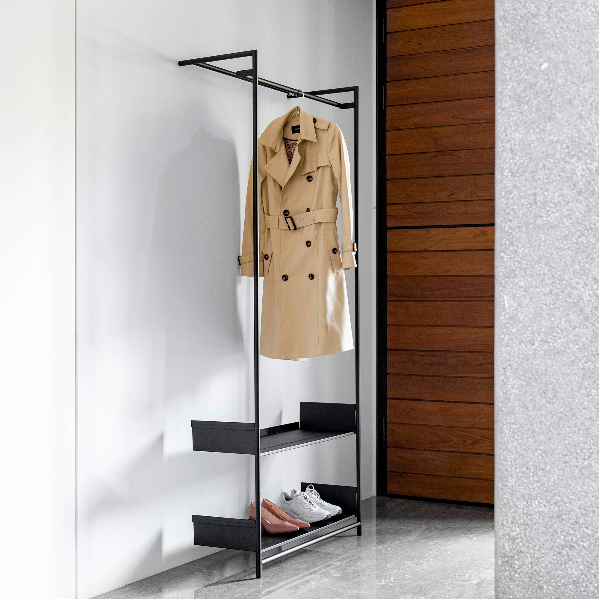 Comba Anlehngarderobe von Mox mit schwarzem Metallrahmen, beigem Trenchcoat und Schuhen auf Ablageflächen.