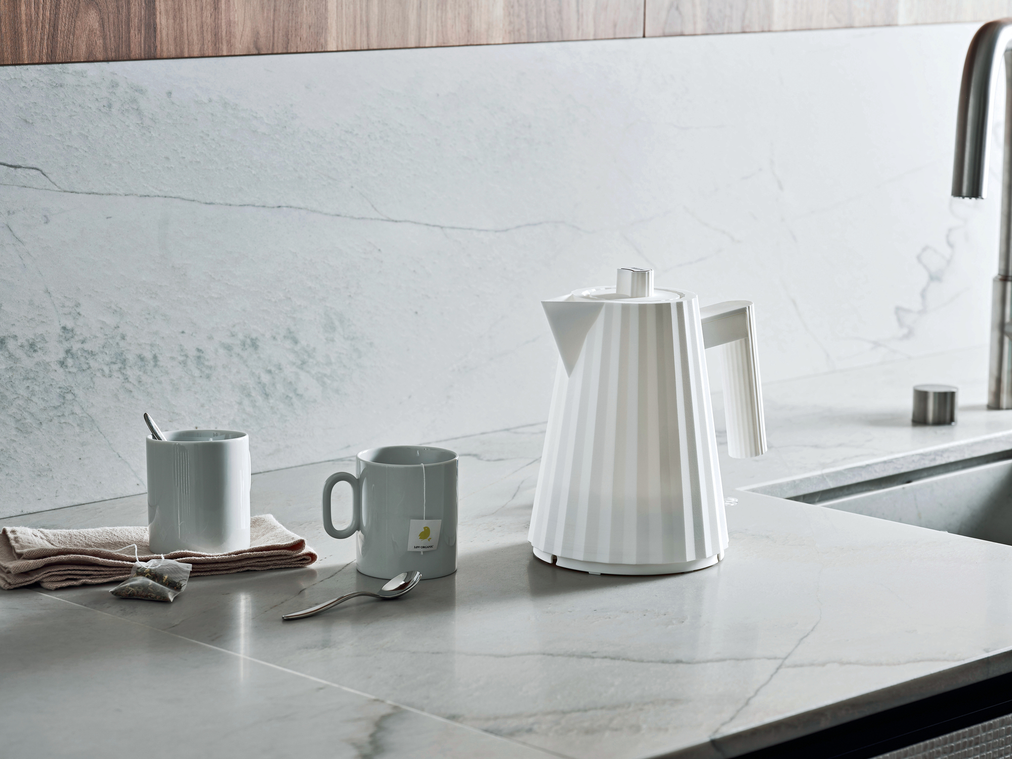 Eleganter, weißer Alessi Plissée Wasserkocher neben Tassen und Teebeuteln auf einer Marmor-Küchenarbeitsplatte.