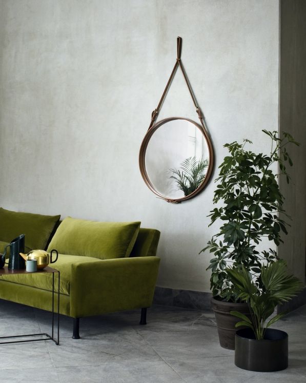 Wohnzimmer mit grünem Samtsofa, Pflanzen und rundem Wandspiegel mit Lederriemen als Dekoration.