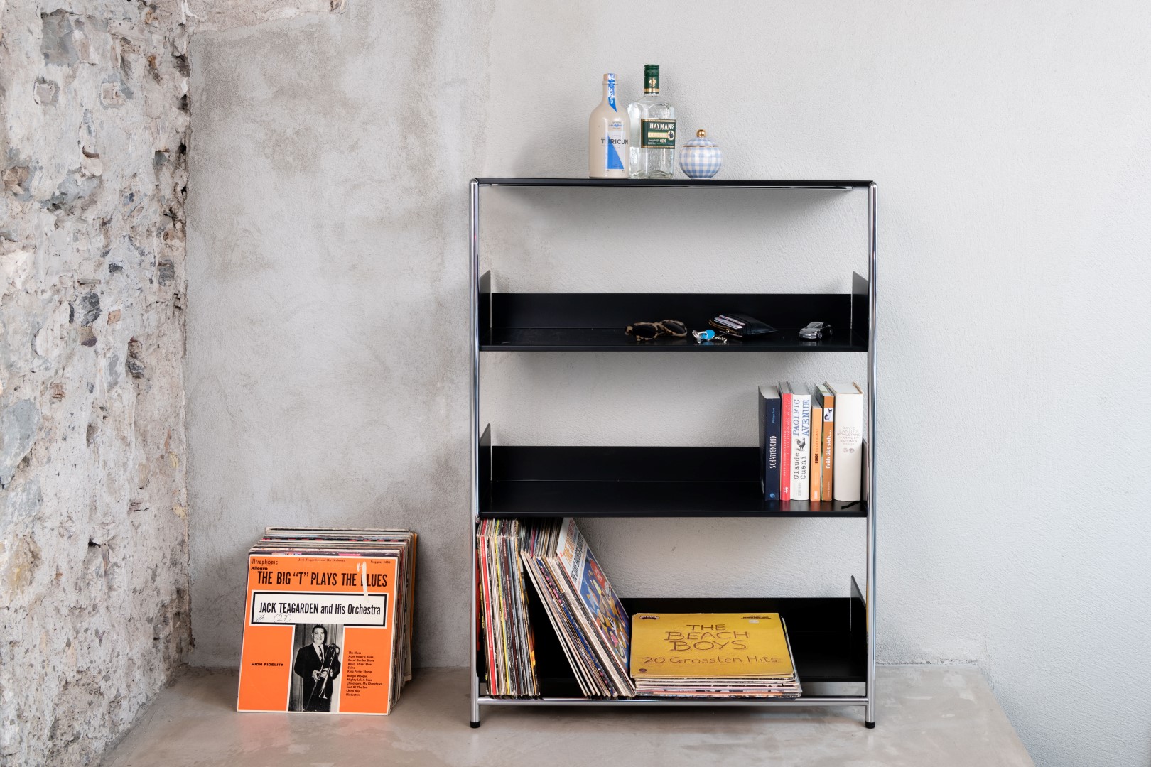 Schwarzes Pool 110 Anlehnregal von Mox mit Schallplatten, Büchern und Dekoration vor einer Betonwand.