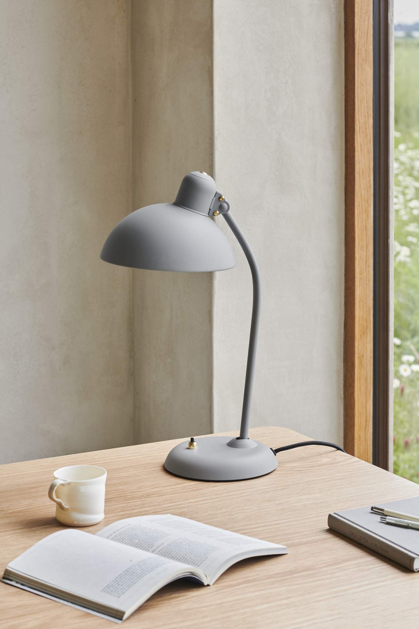 Graue KAISER idell Tischleuchte auf Schreibtisch mit Buch, Tasse und Notizbuch. Designlampe für Büro.