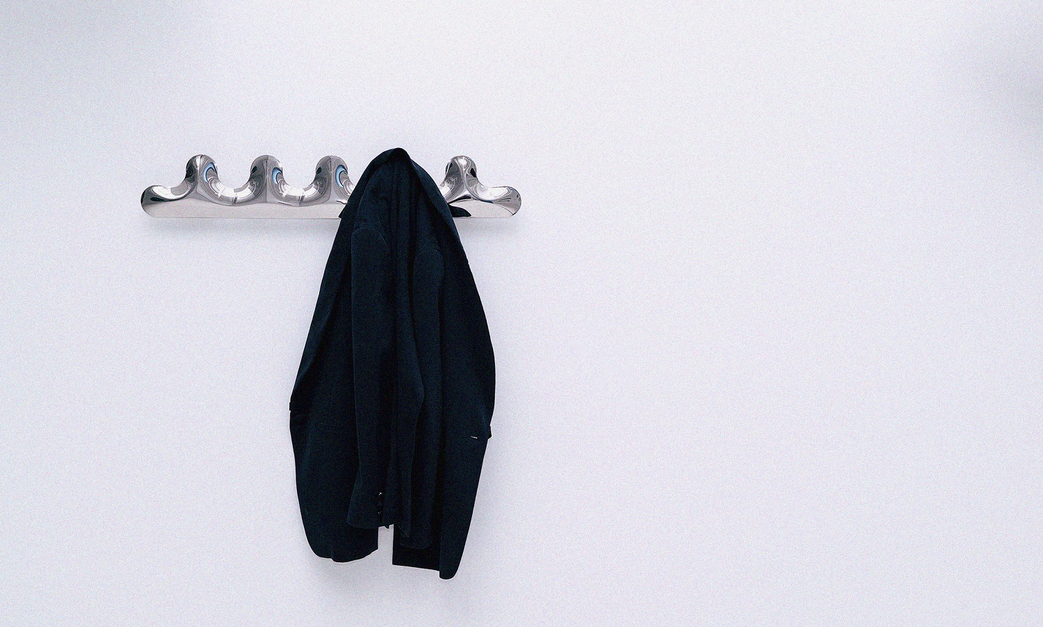 Kamm Garderobe Zieta aus poliertem Stahl an weißer Wand mit hängendem, dunklem Mantel.