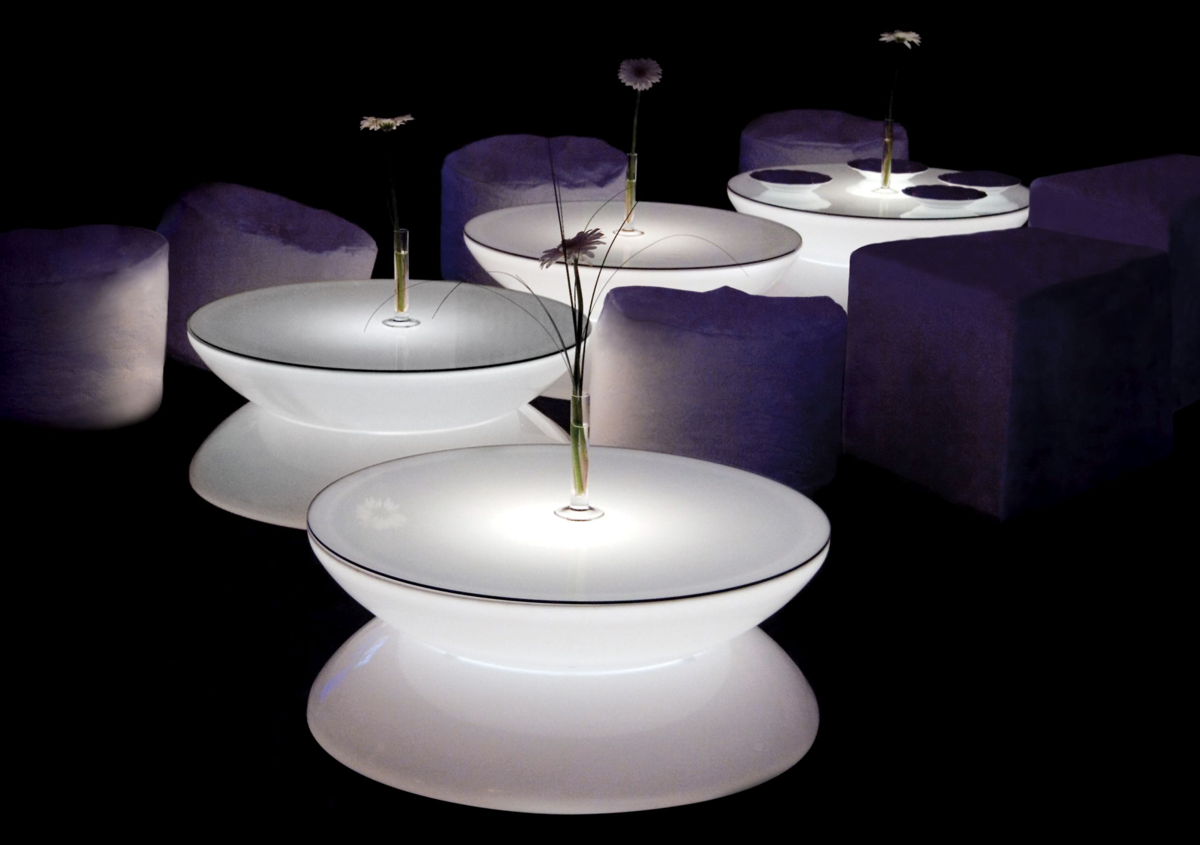 Moree Lounge Tisch Indoor: Runde, beleuchtete Tische und lilafarbene Sitzhocker in dunklem Raum.