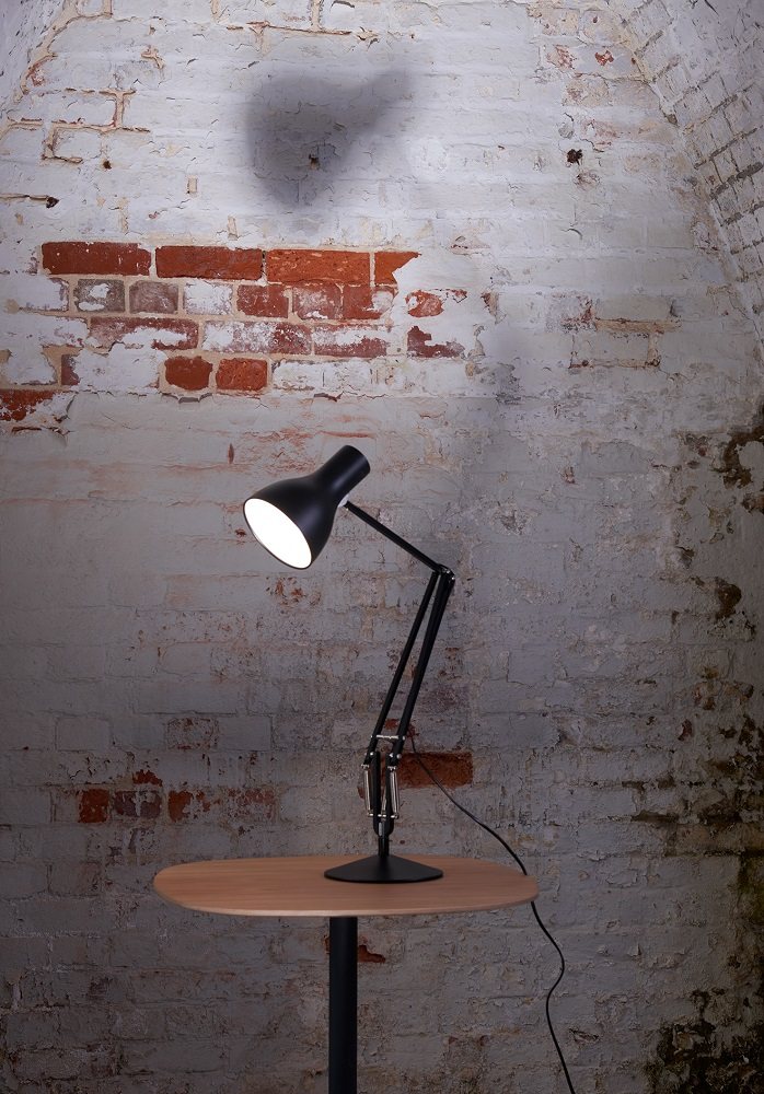 Schwarze Anglepoise Type 75 Tischleuchte auf einem Holztisch vor einer Backsteinwand, modernes Design für Büro und Zuhause.