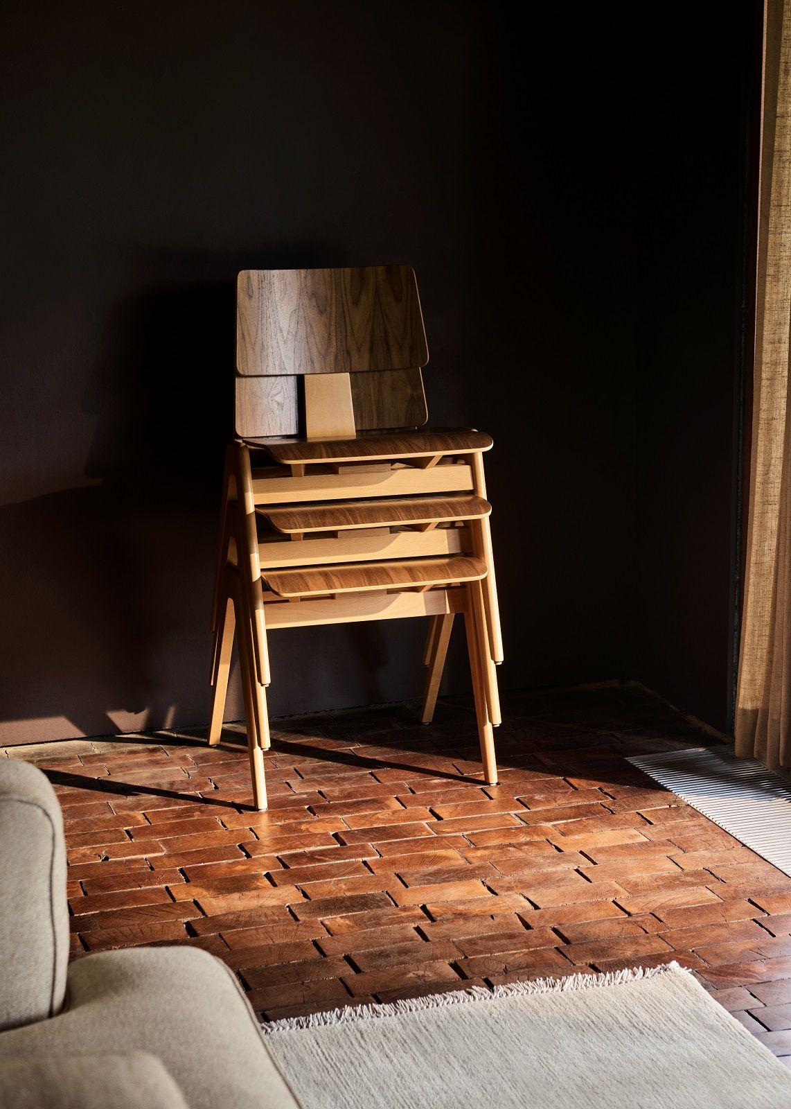 Gestapelte AndTradition Daystak RD1 Esszimmerstühle aus Holz auf einem Holzboden vor dunkler Wand.